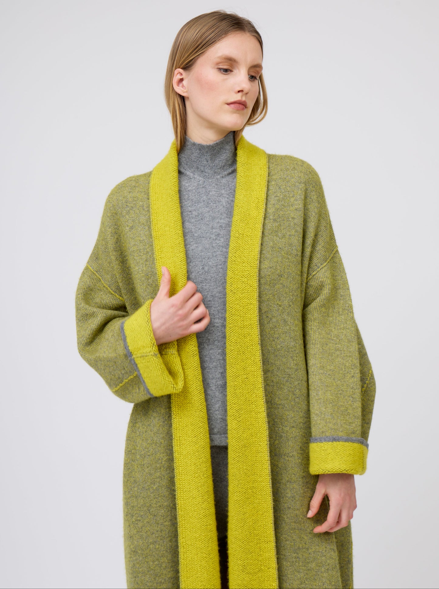 Close-up model in long mustard yellow and grey mix cashmere knit coat with yellow collar revere and turn back cuffs; wearing with grey turtle neck knit top.