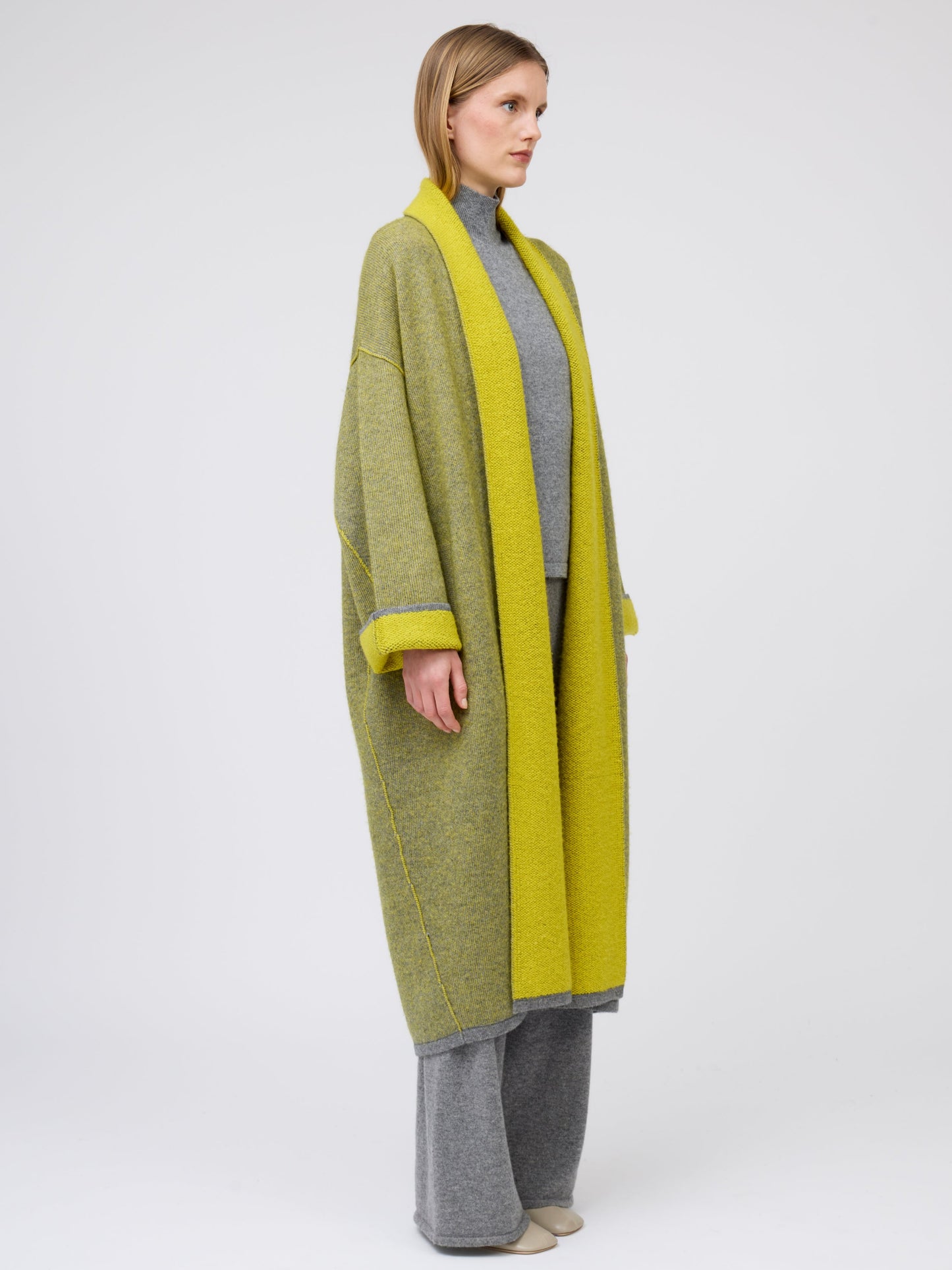 Side view model in long mustard yellow and grey mix cashmere knit coat with yellow collar revere and turn back cuffs; wearing with grey knit turtle neck top and wide leg cashmere pants.