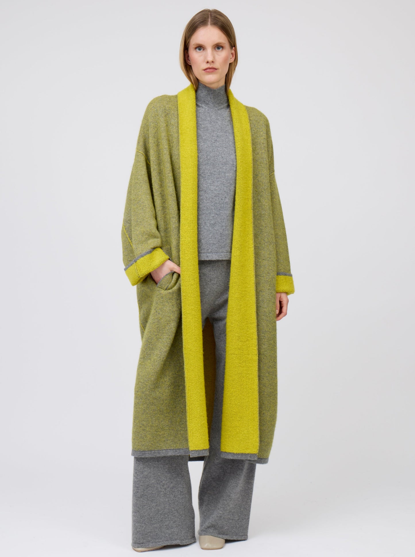 Front view model in long length mustard yellow and grey mix cashmere knit coat with yellow collar revere, turn back cuffs and pockets; wearing with grey knit turtle neck top and wide leg cashmere pants.