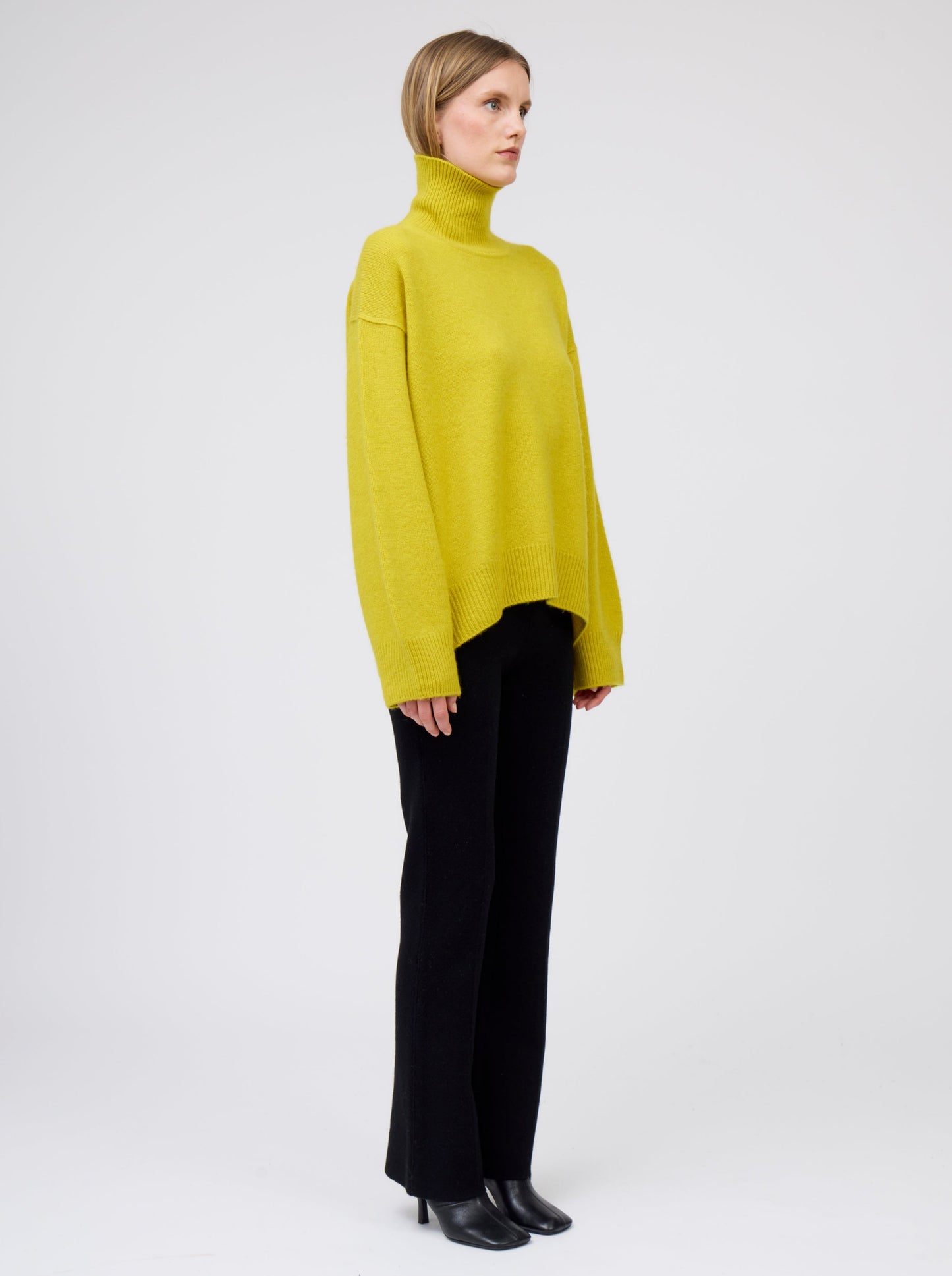 Side view model wears mustard yellow cashmere roll neck oversized jumper with black tailored cashmere pants and high heeled boots.