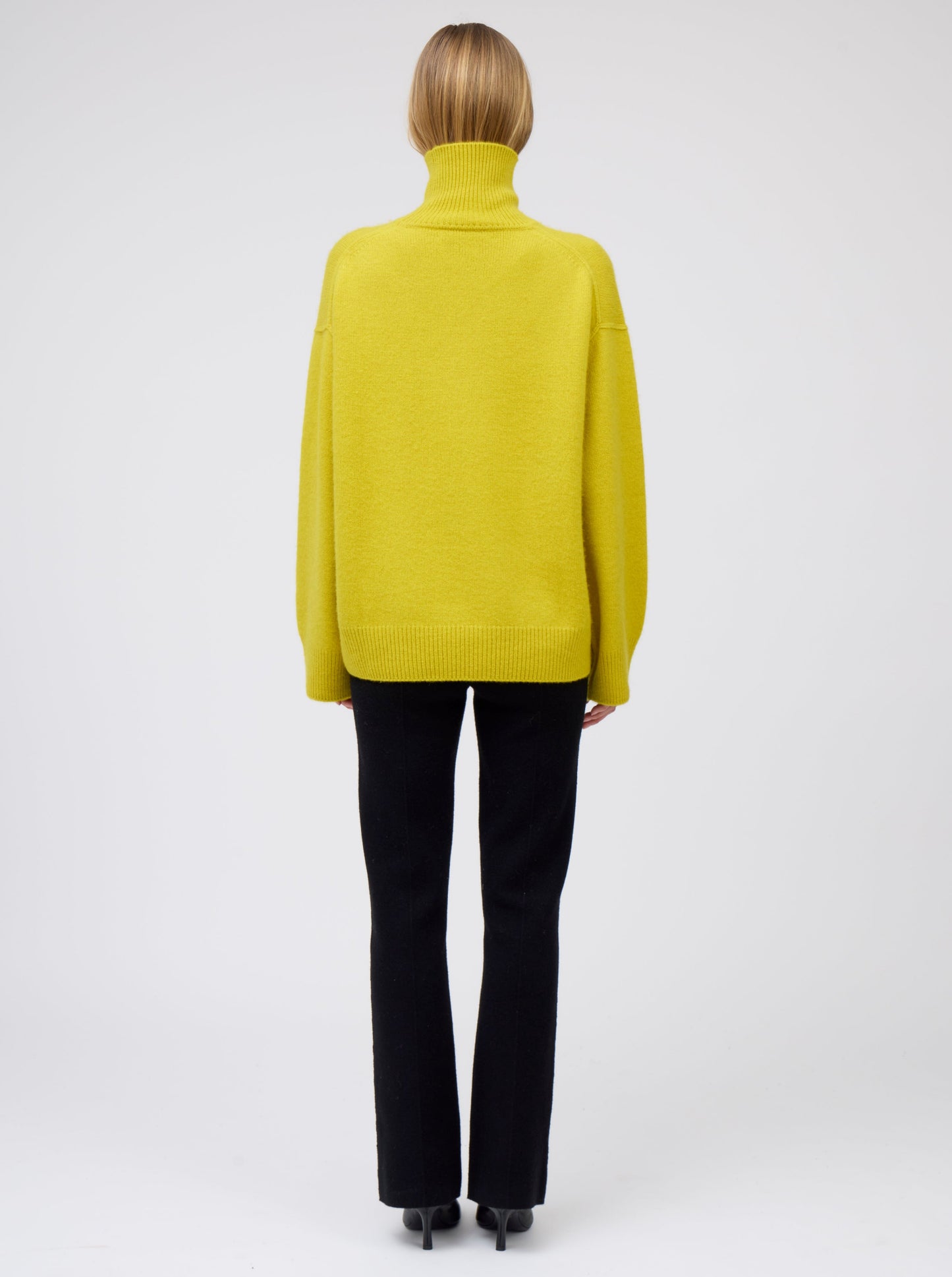 Back view model wears mustard yellow cashmere roll neck oversized jumper with black tailored cashmere pants and high heeled boots.