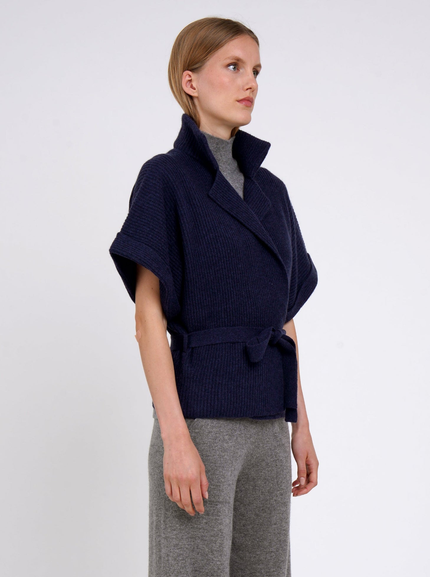 Cropped side view model wears navy cashmere short ribbed knit jacket over grey knit top and grey wide leg pants.