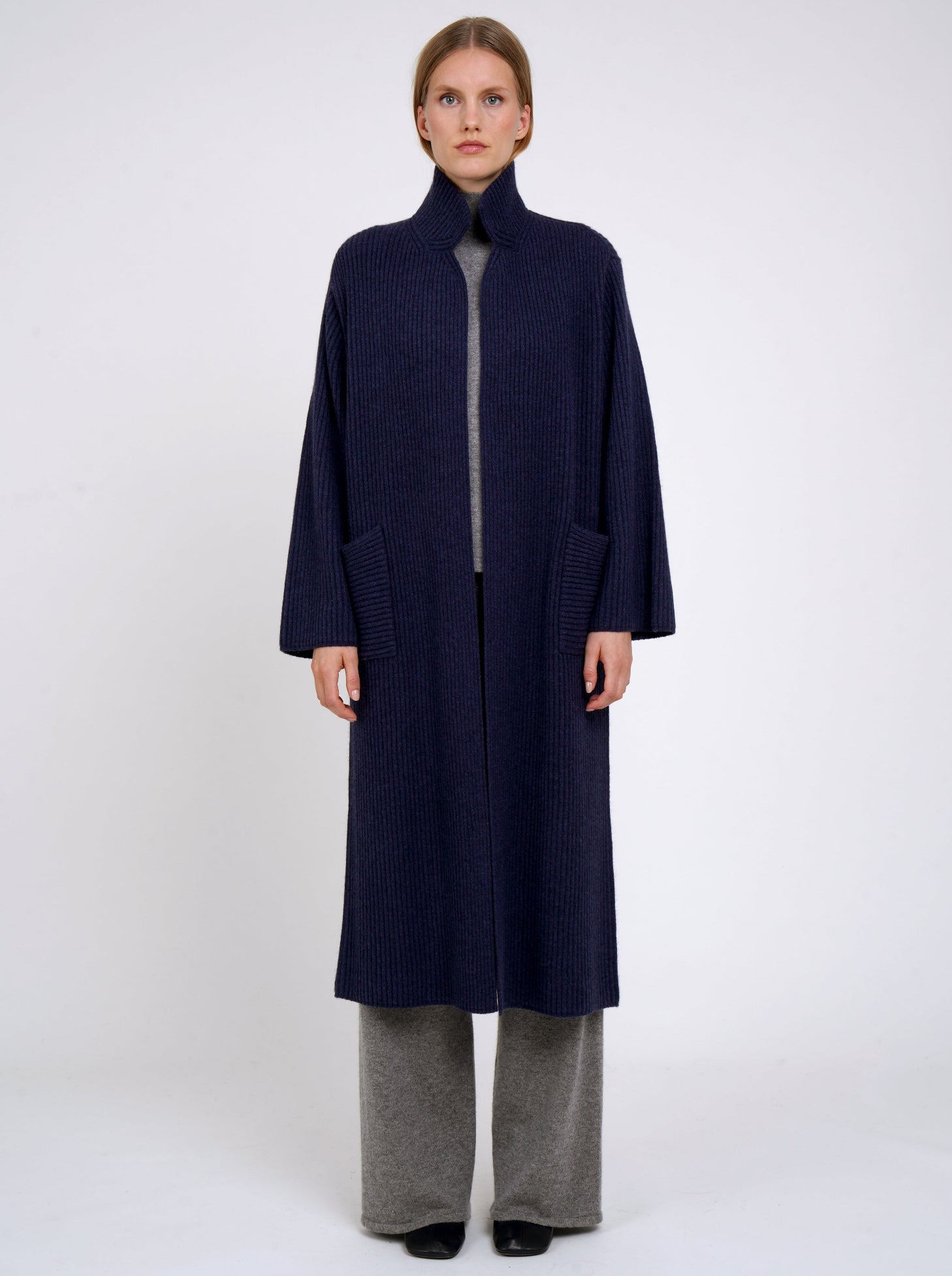 Model wearing a long navy cardigan coat in ribbed knit cashmere with high collar, patch pockets and generous long sleeves; worn with grey cashmere pants.