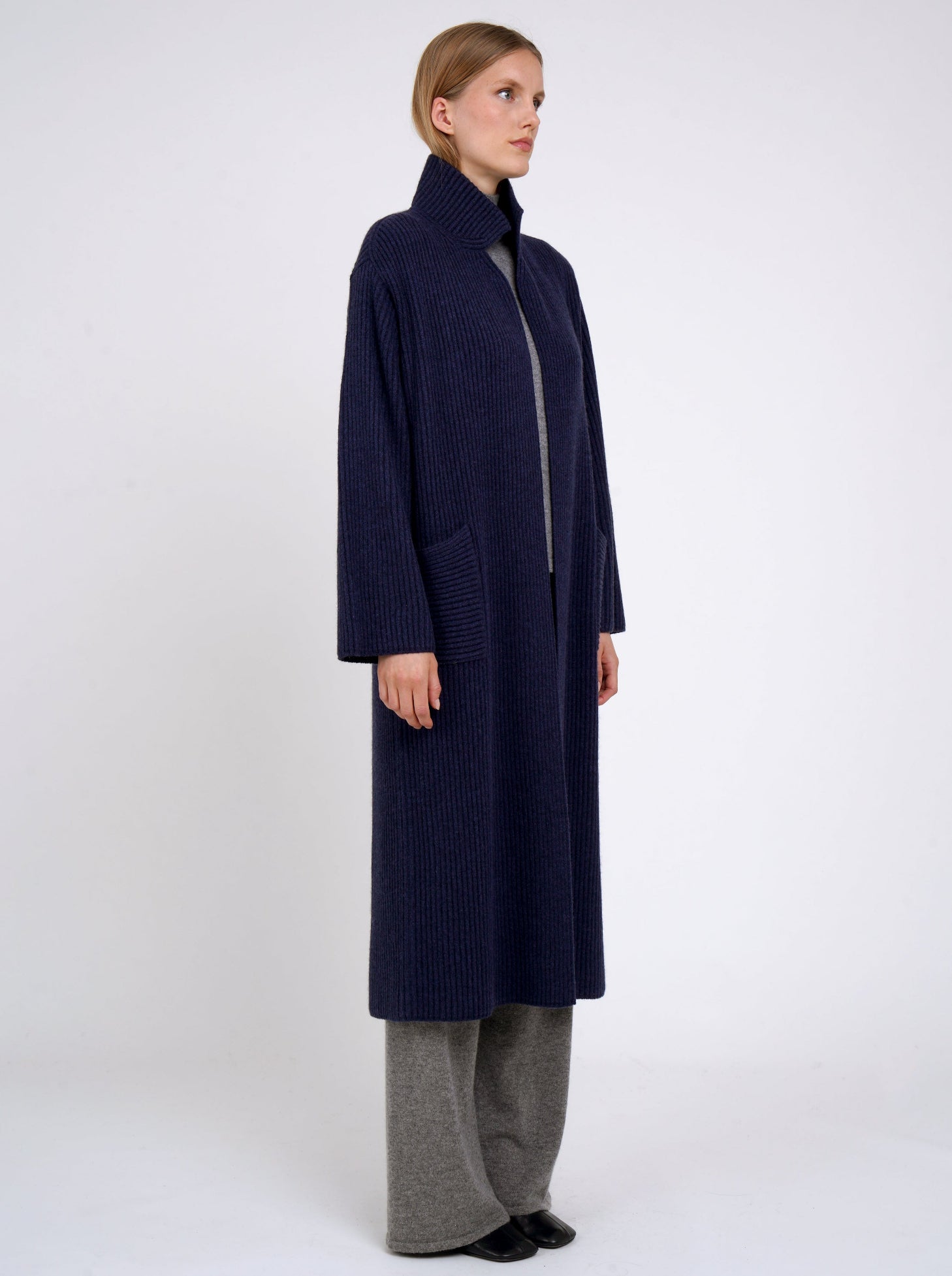 Side view model wearing a long navy cardigan coat in ribbed knit cashmere with high collar, patch pockets and generous long sleeves; worn with grey cashmere pants.
