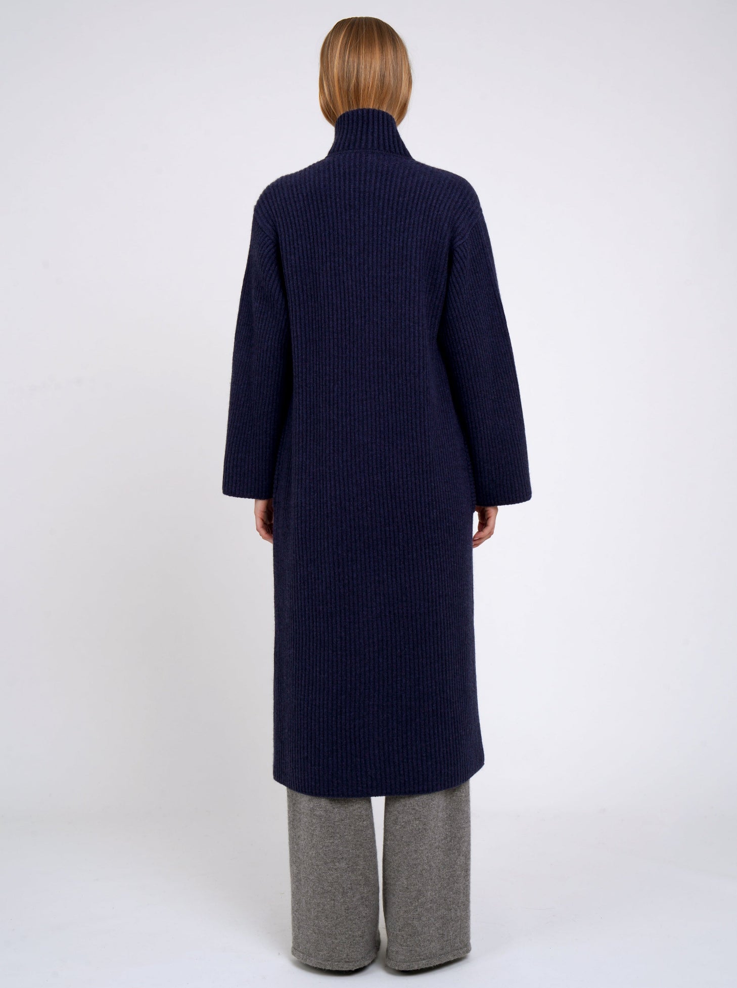 Back of model wearing a long navy coat in ribbed knit cashmere with high collar, patch pockets and generous long sleeves.