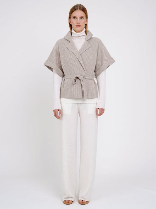 Front view model wears beige cashmere short ribbed knit jacket with short sleeves, over white, turtle neck, long sleeve top and white wide leg pants.