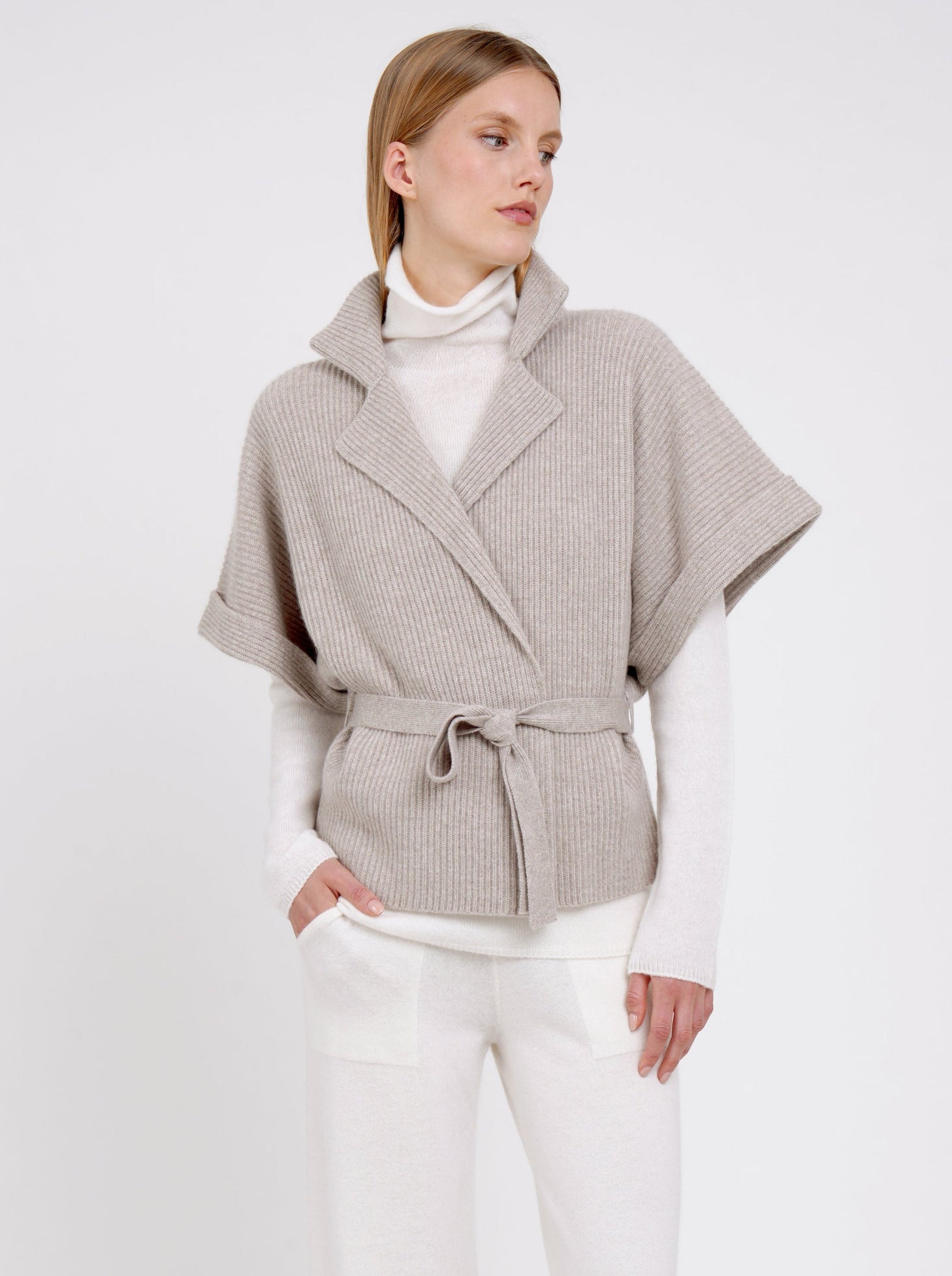 Close up image model wearing beige cashmere short-sleeved ribbed knit short jacket over white knit top and white wide leg pants.