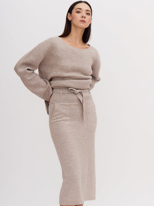 Model wearing outfit in beige cashmere of high waisted pencil skirt with tie belt and matching low neck jumper.