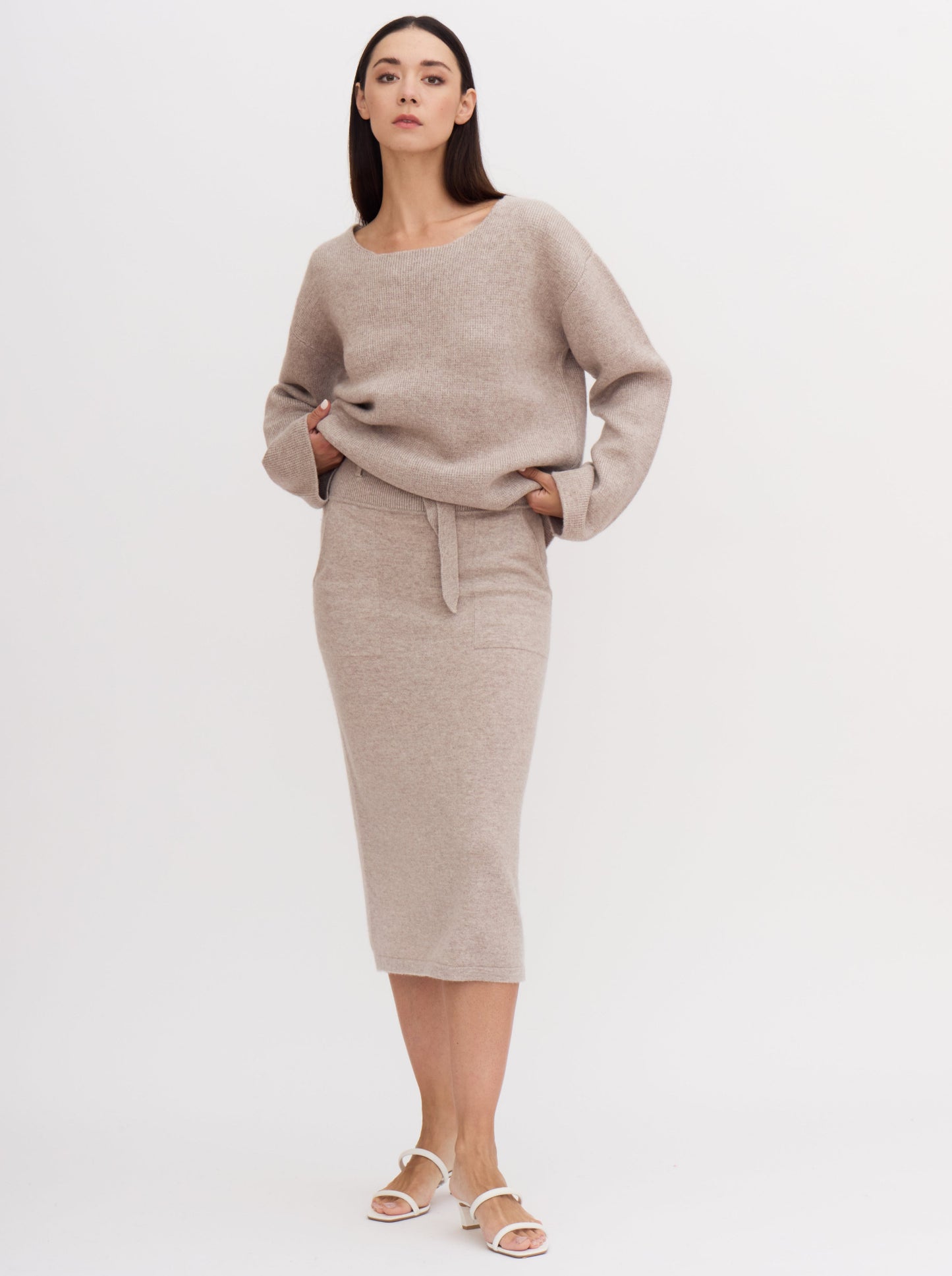 Model wearing outfit in beige cashmere of high waisted, calf length pencil skirt  and matching low neck jumper with wide sleeves.