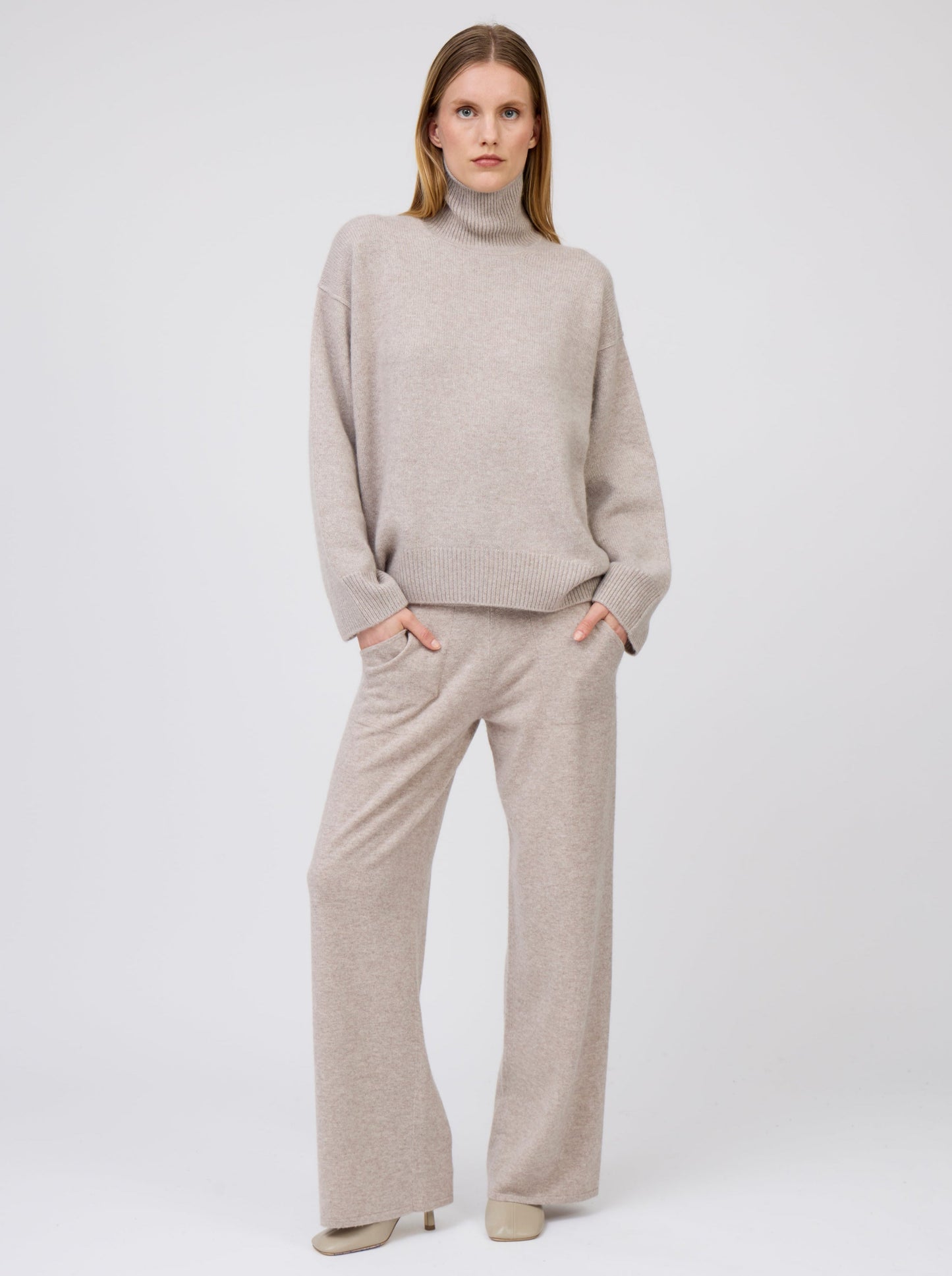 Model wears beige cashmere, wide leg pants with pockets; worn with matching beige cashmere oversized roll neck jumper with long sleeves.
