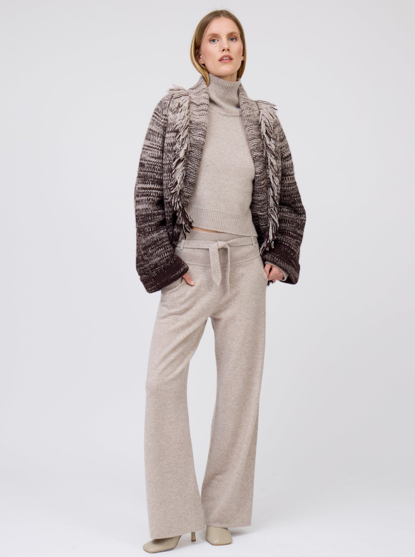 Front view of model wearing beige cashmere, belted, wide leg pants with pockets; worn with knitted brown and beige mix short jacket with fringe collar over roll neck beige sweater.