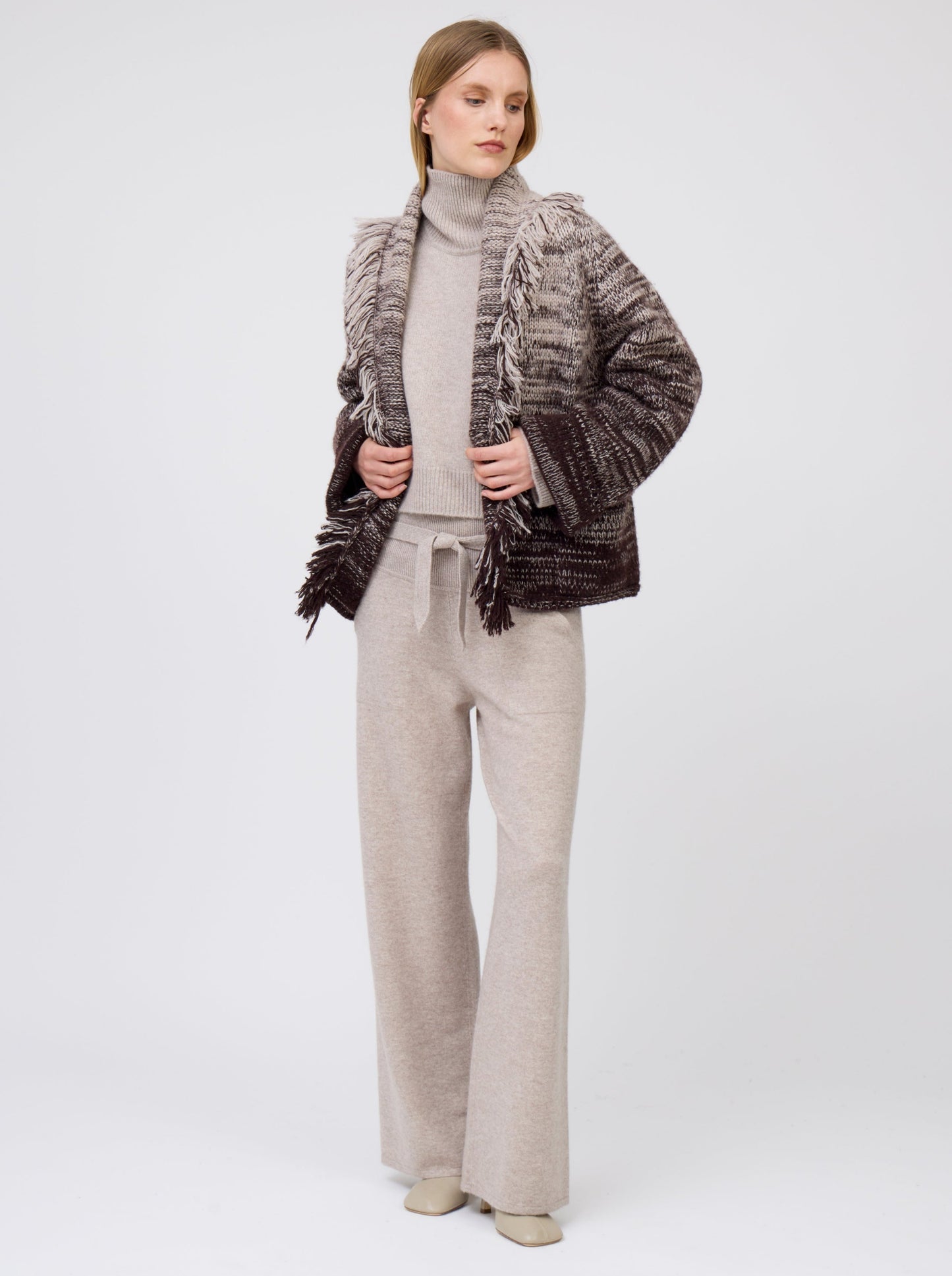 Model wears beige cashmere, belted, wide leg pants with pockets; worn with knitted brown and beige mix short jacket with fringe collar over cropped, roll neck, beige sweater.