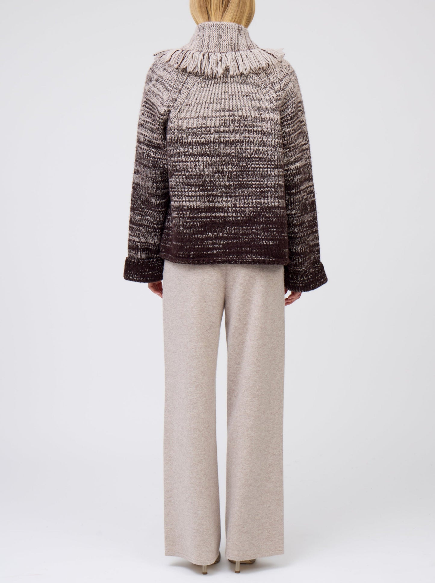 Back view of model wearing beige cashmere wide leg pants; worn with knitted brown and beige mix short jacket with fringed shawl collar and wide sleeves.
