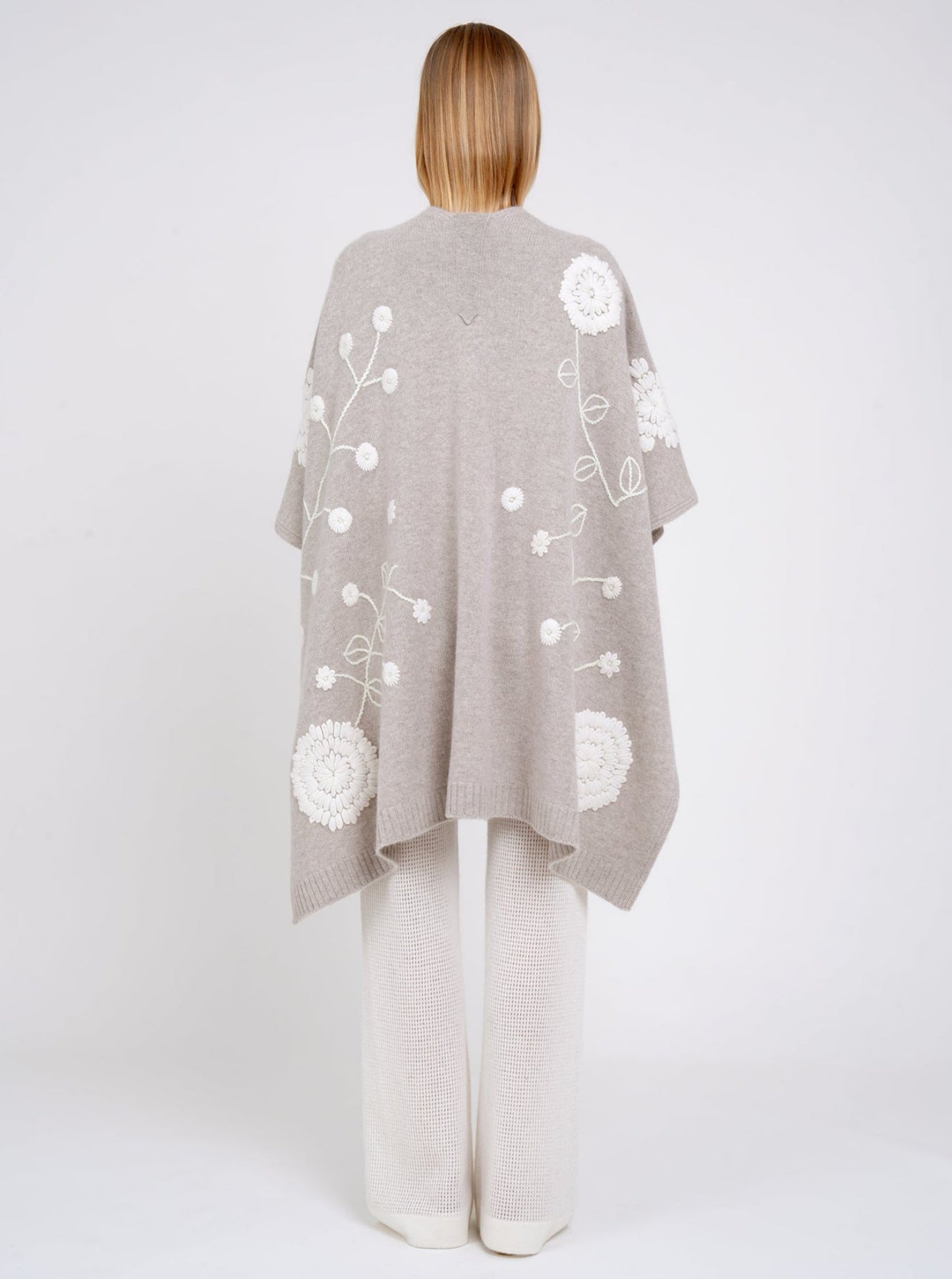 Back view model in beige cashmere long shawl with large white embroidered flowers over white knit pants.