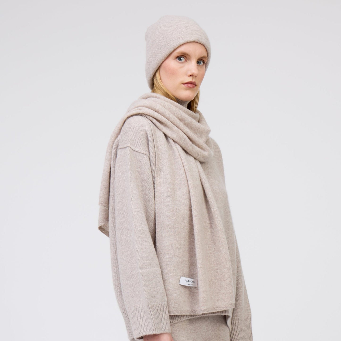 Cropped side view of model wearing complete outfit in matching beige cashmere: beanie hat, scarf and oversized jumper.