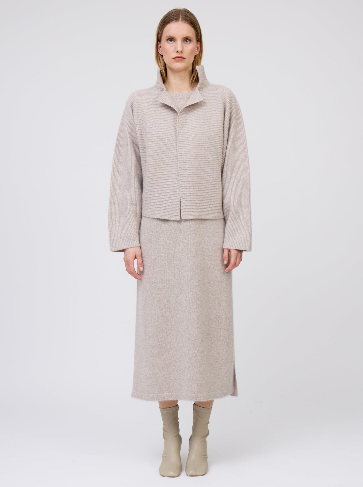 Front view of model wearing beige cashmere, ribbed knit, short jacket with high collar and kimono sleeves over matching beige cashmere straight cut dress and beige boots.