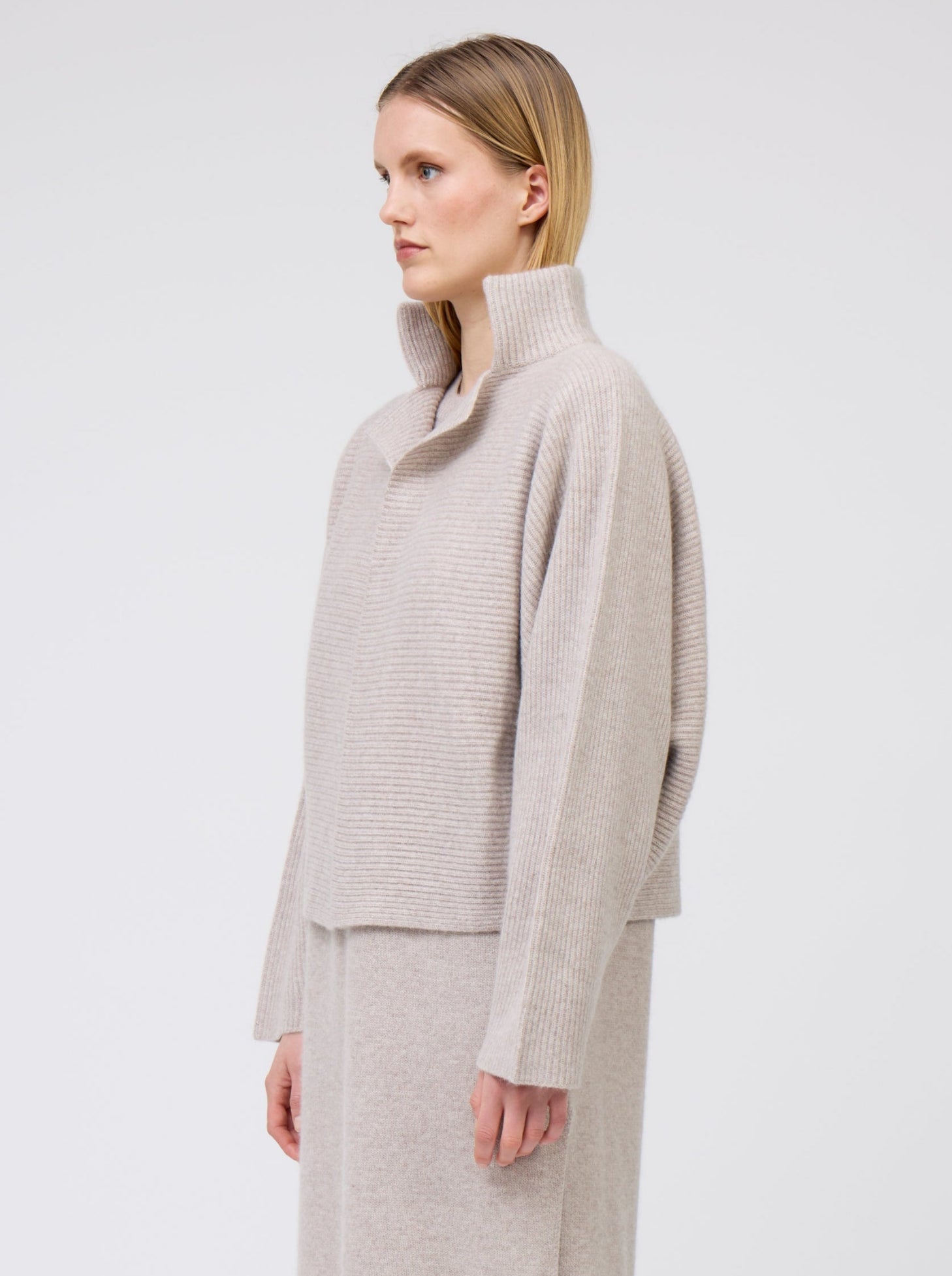 Close-up side view of model wearing beige cashmere, ribbed knit, short jacket with high collar and kimono sleeves.