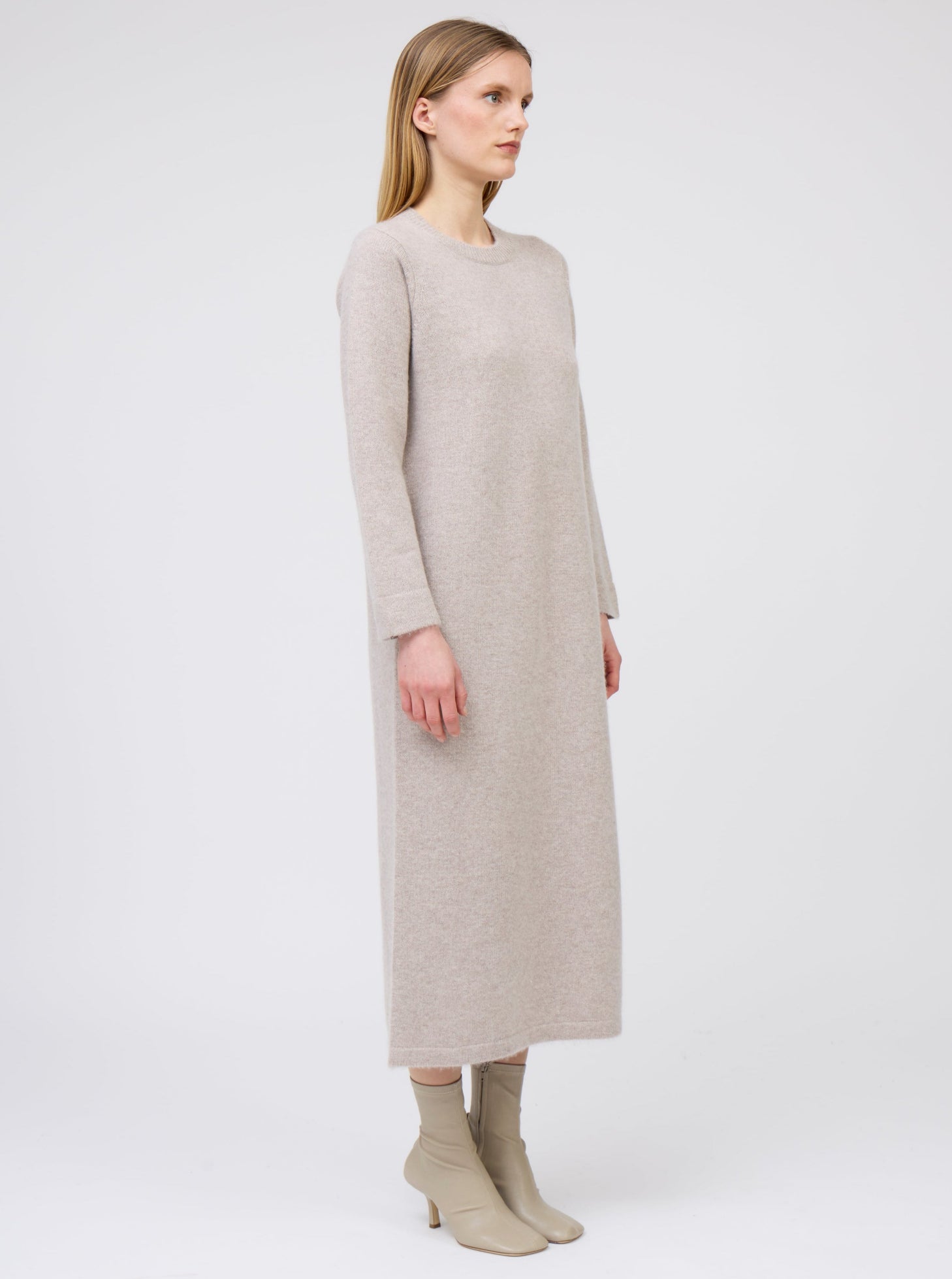 Side view of model wearing plain round neck maxi dress in beige cashmere, with long sleeves and side slit, worn with heeled beige boots.