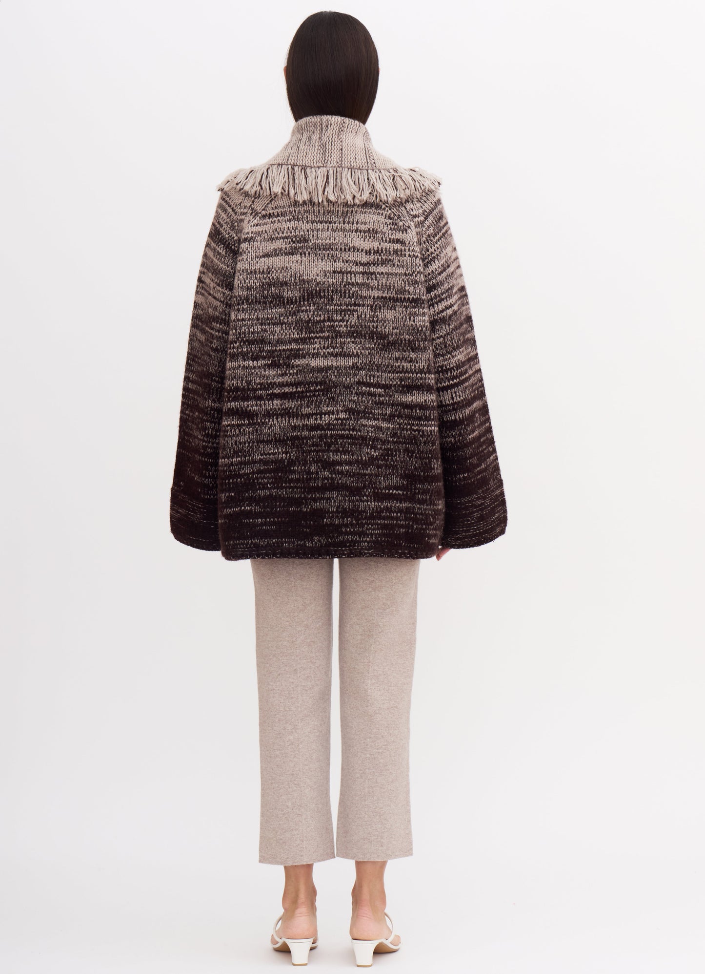 Back view of model wearing chunky knit jacket with fringed shawl collar in a cashmere colour mix of beige and dark brown; with beige cashmere cropped pants.
