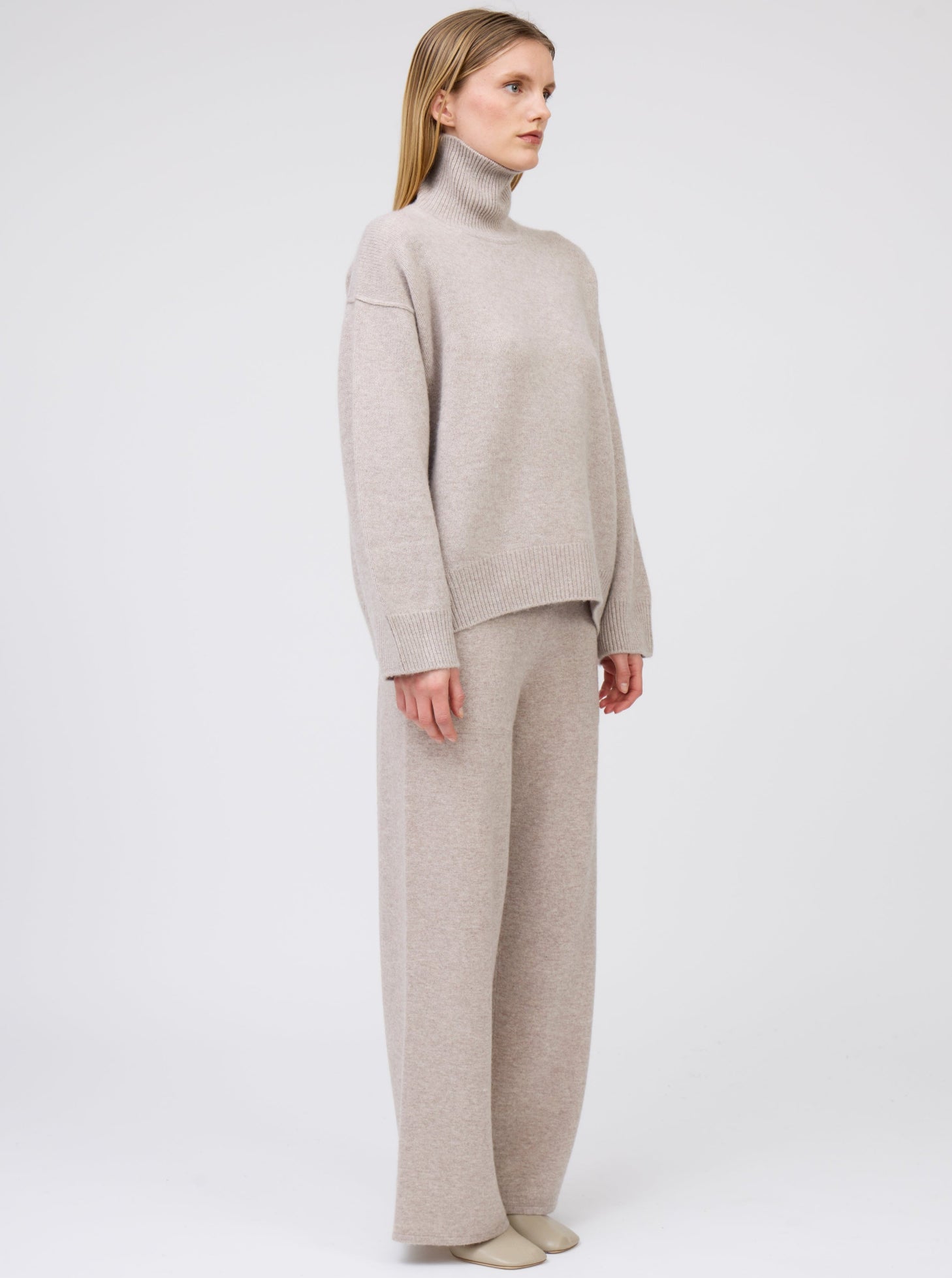 Side view of model wearing beige cashmere oversized roll neck jumper with generous long sleeves and high-low hem; worn with beige cashmere wide leg trousers.