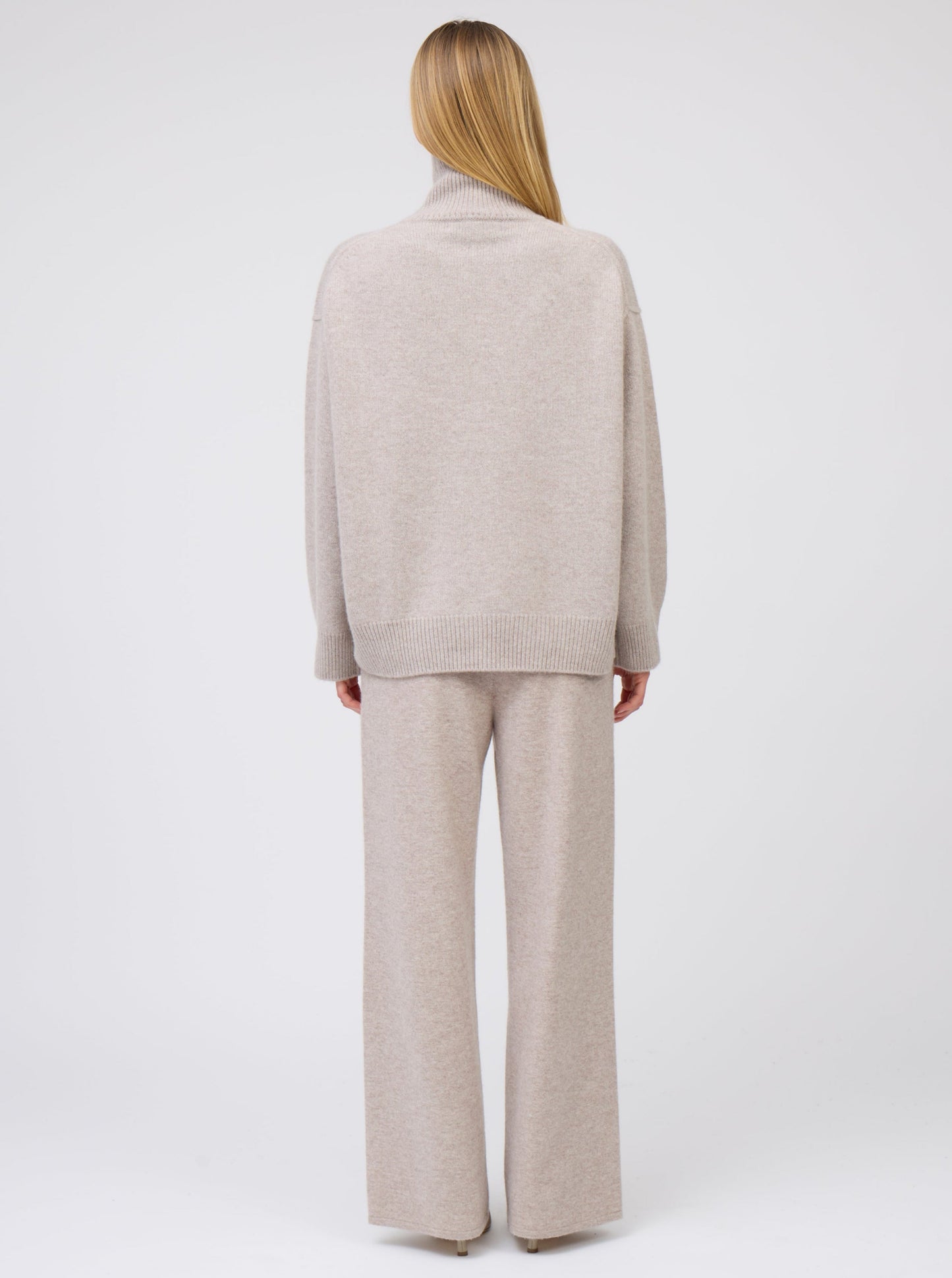 Back view of model wearing beige cashmere oversized roll neck jumper with generous long sleeves; worn with matching beige cashmere wide leg pants.