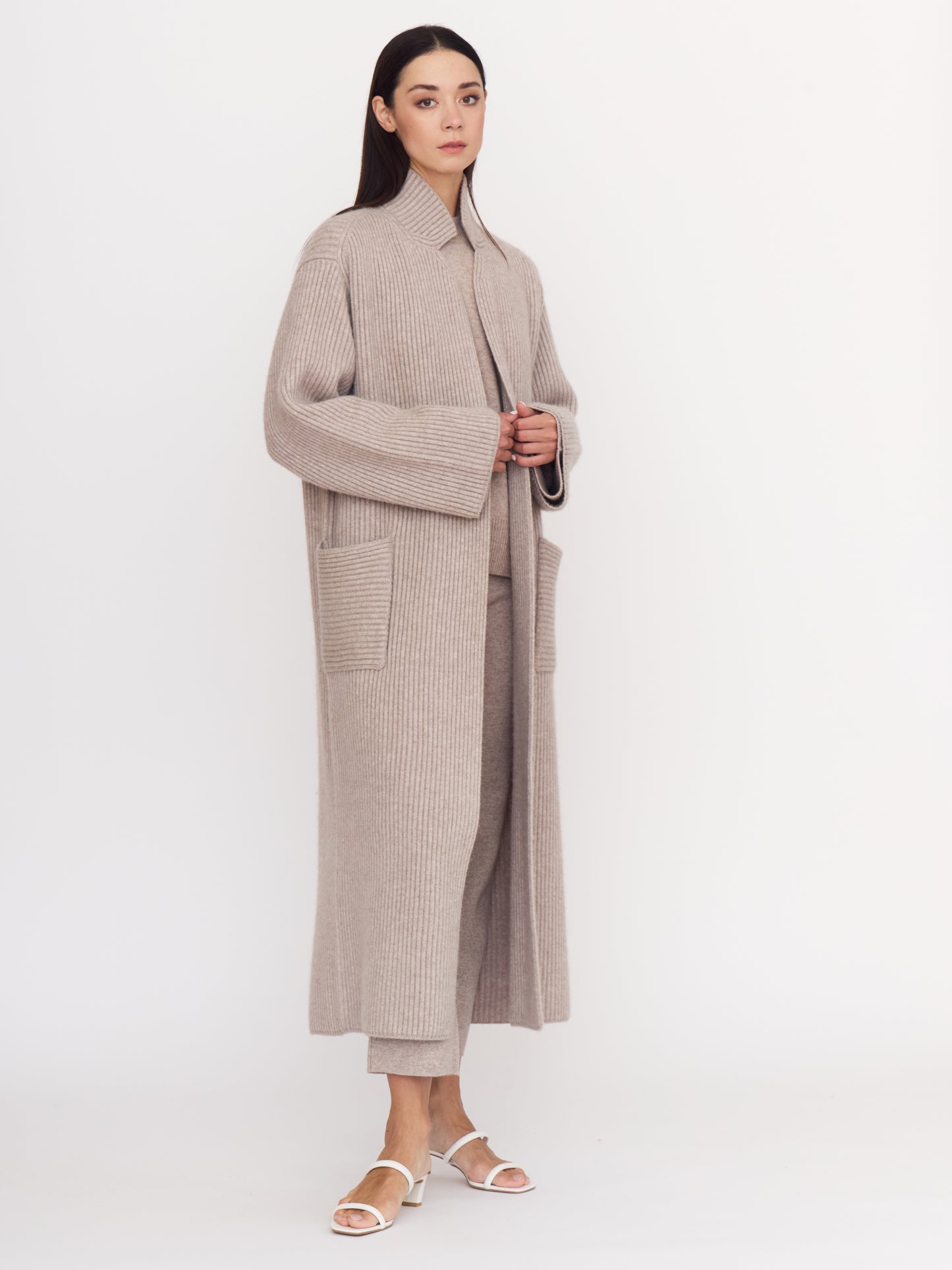Side angle view of model wearing cool beige shade of ribbed cashmere knit long coat with long sleeves and patch pockets.