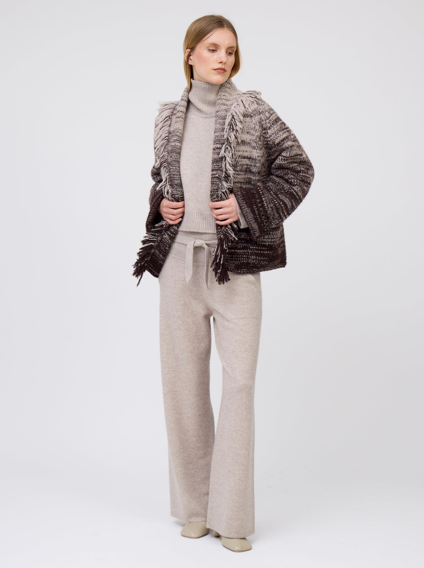 Front view of model wearing colour mix knitted short jacket with fringe collar in beige and dark brown cashmere over roll neck beige sweater and wide leg cashmere pants.