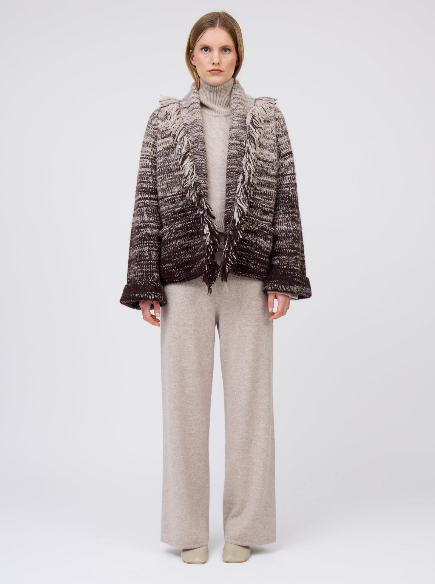 Model wears  gradient colour mix knitted short jacket in beige and dark brown over roll neck beige sweater and wide leg pants.