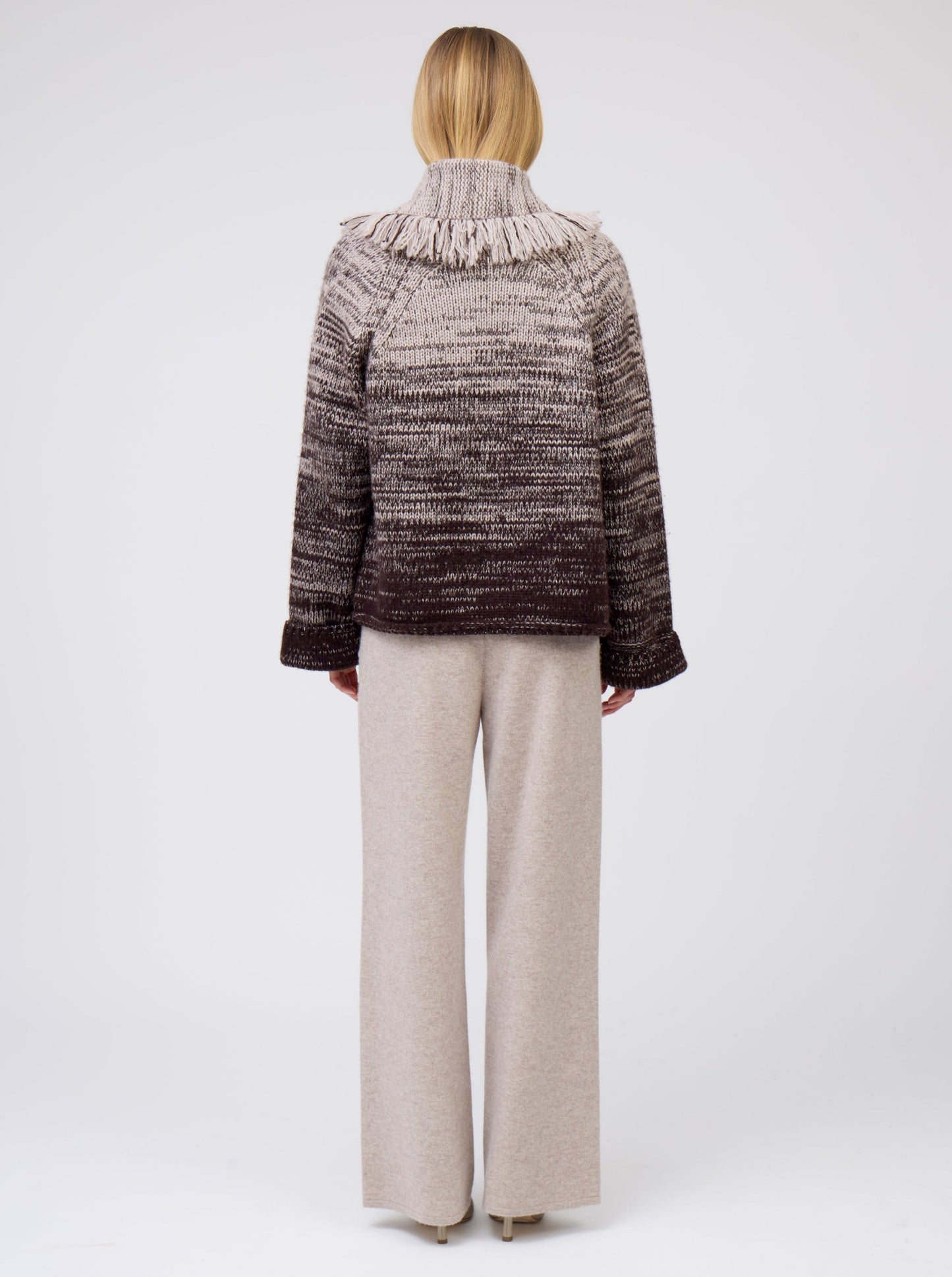 Back view of model wearing two colour mix knitted cashmere short jacket with fringe collar in beige and dark brown with matching beige cashmere wide leg pants.