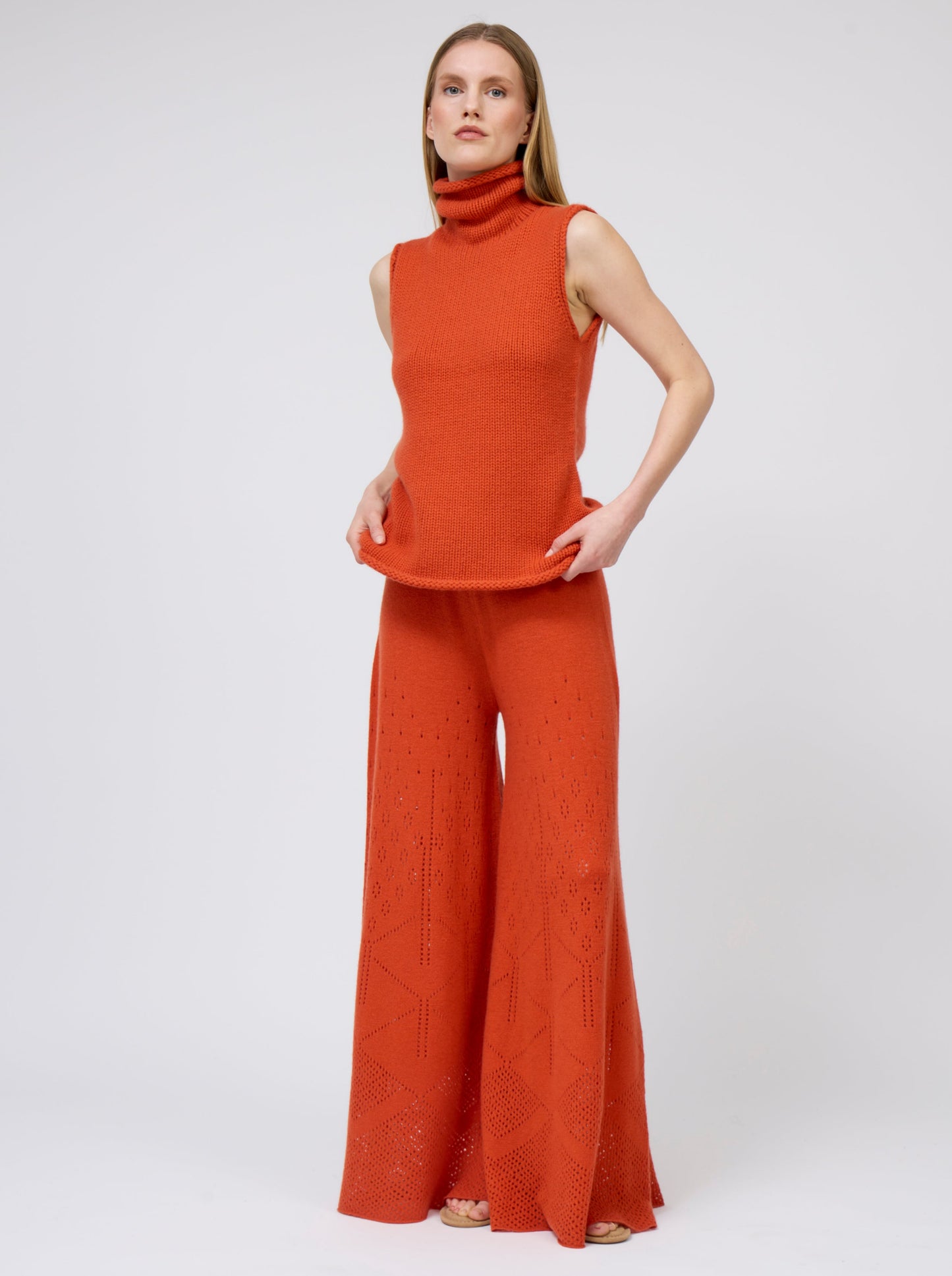 Front view model wears bright orange sleeveless chunky cashmere knit roll neck jumper with matching orange pants.