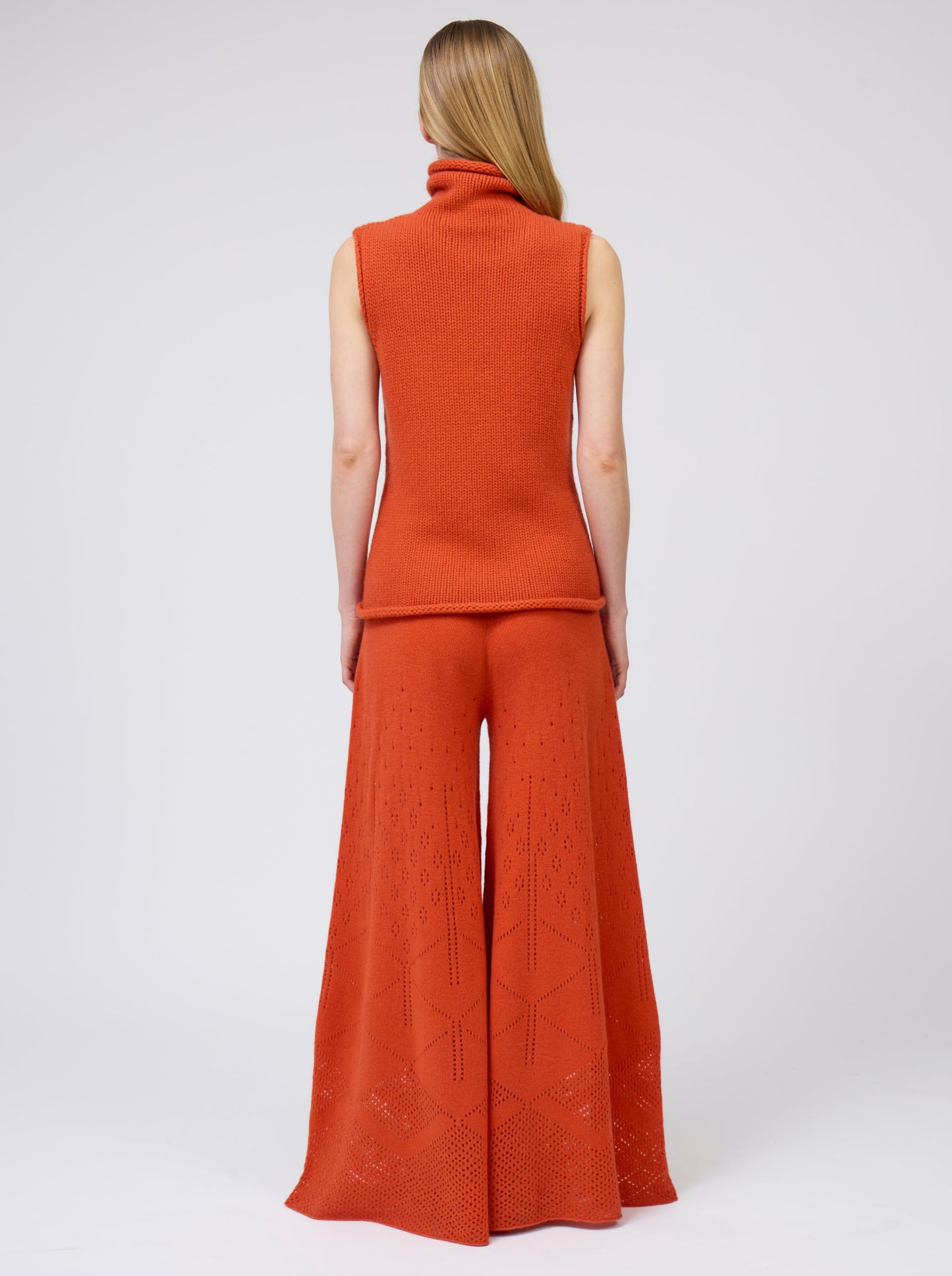 Back view model wears bright orange sleeveless chunky cashmere knit roll neck jumper with very flared cut, matching orange pants.