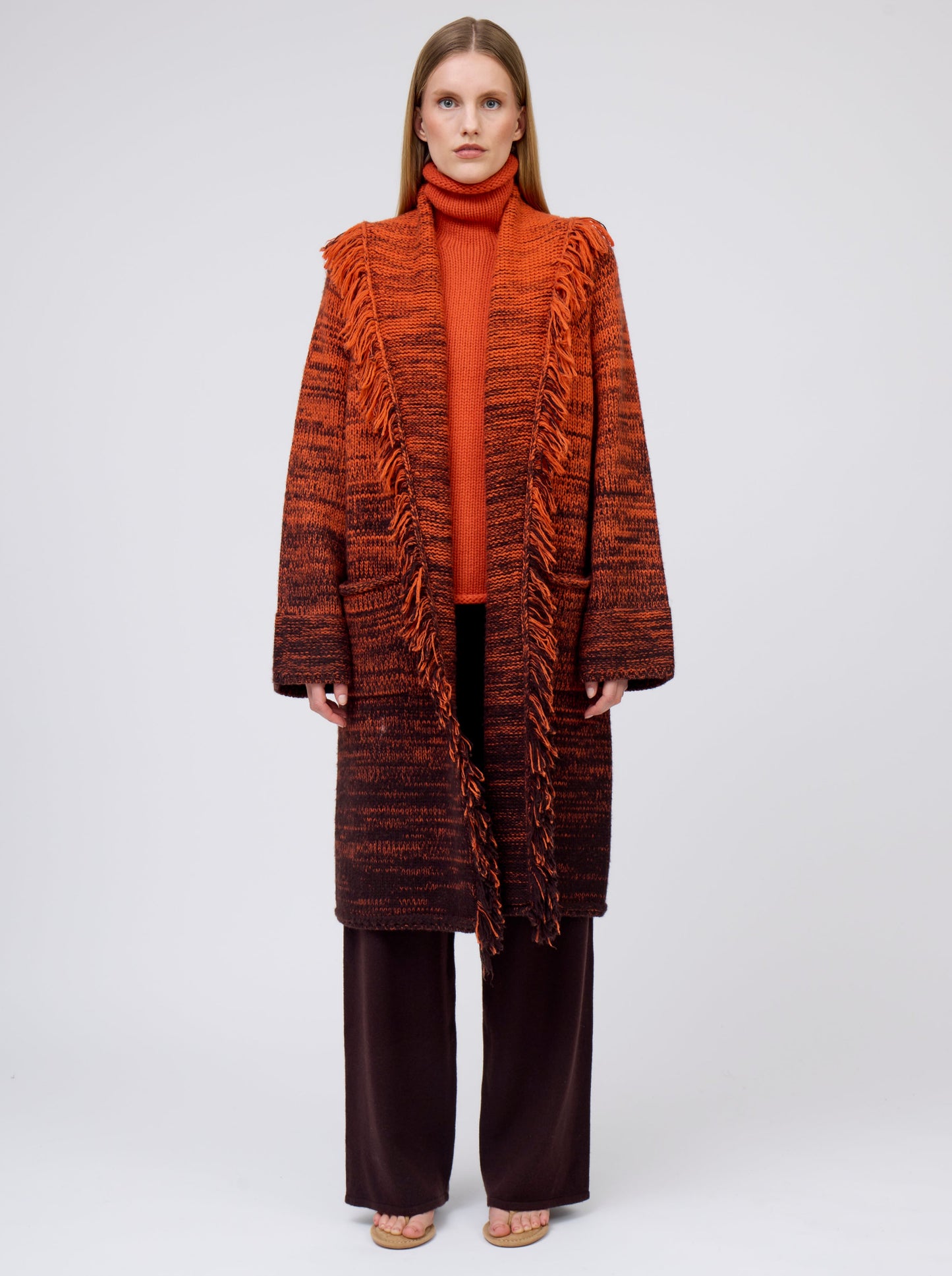 Front view model wearing orange/black mix knit cashmere coat with fringe trim shawl collar; worn with orange jumper and brown pants.