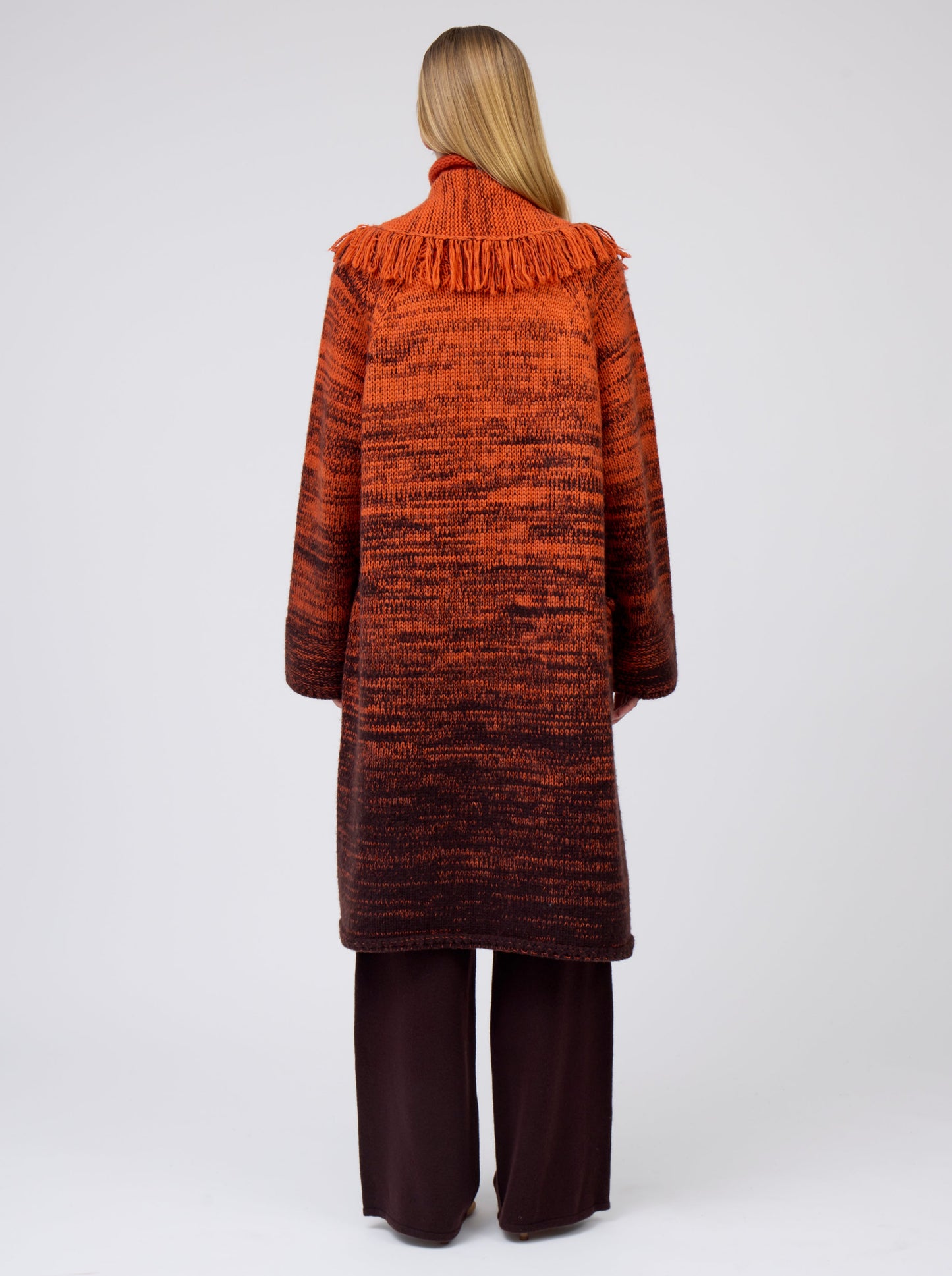Back view model wearing orange/black mix knit cashmere coat with fringe trim shawl collar; worn with brown pants.