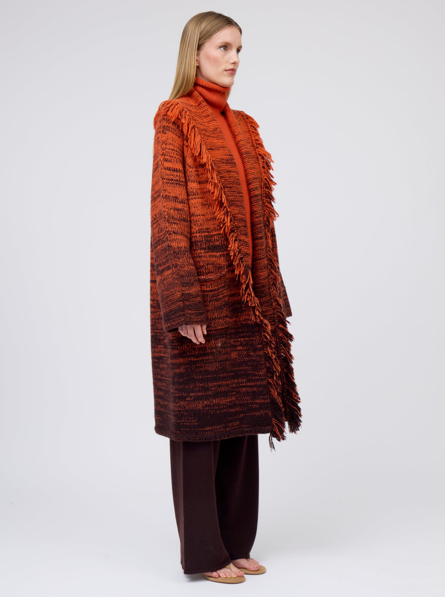 Side view model wearing orange/black mix knit cashmere coat with fringe trim shawl collar; worn with orange jumper and brown pants.