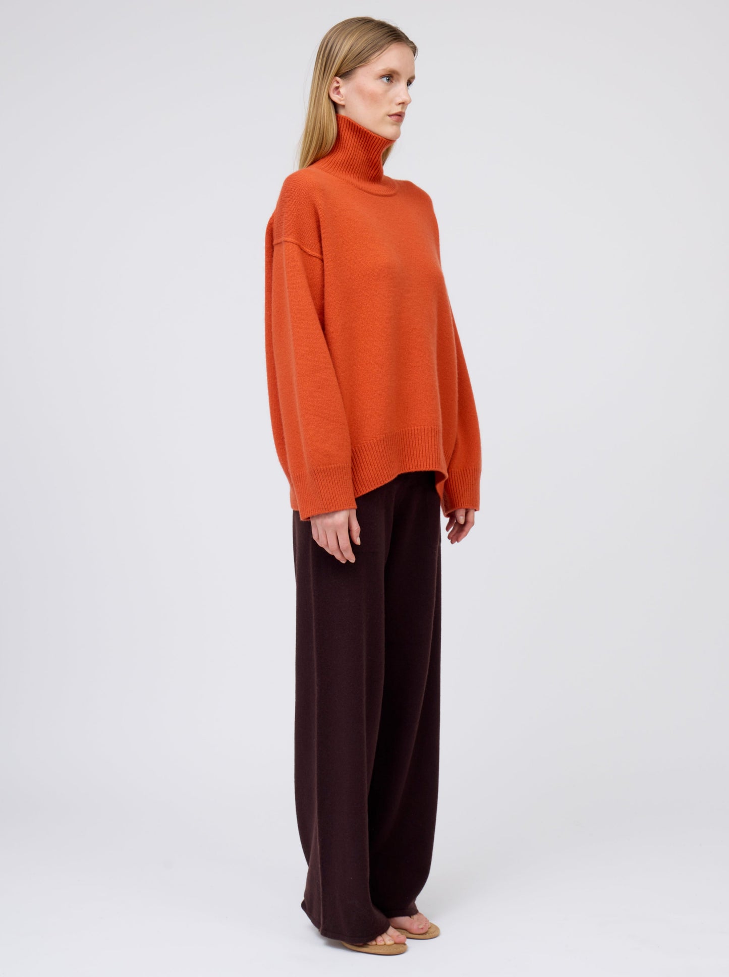 Side view model wears deep orange cashmere roll neck jumper with wide leg dark brown pants.