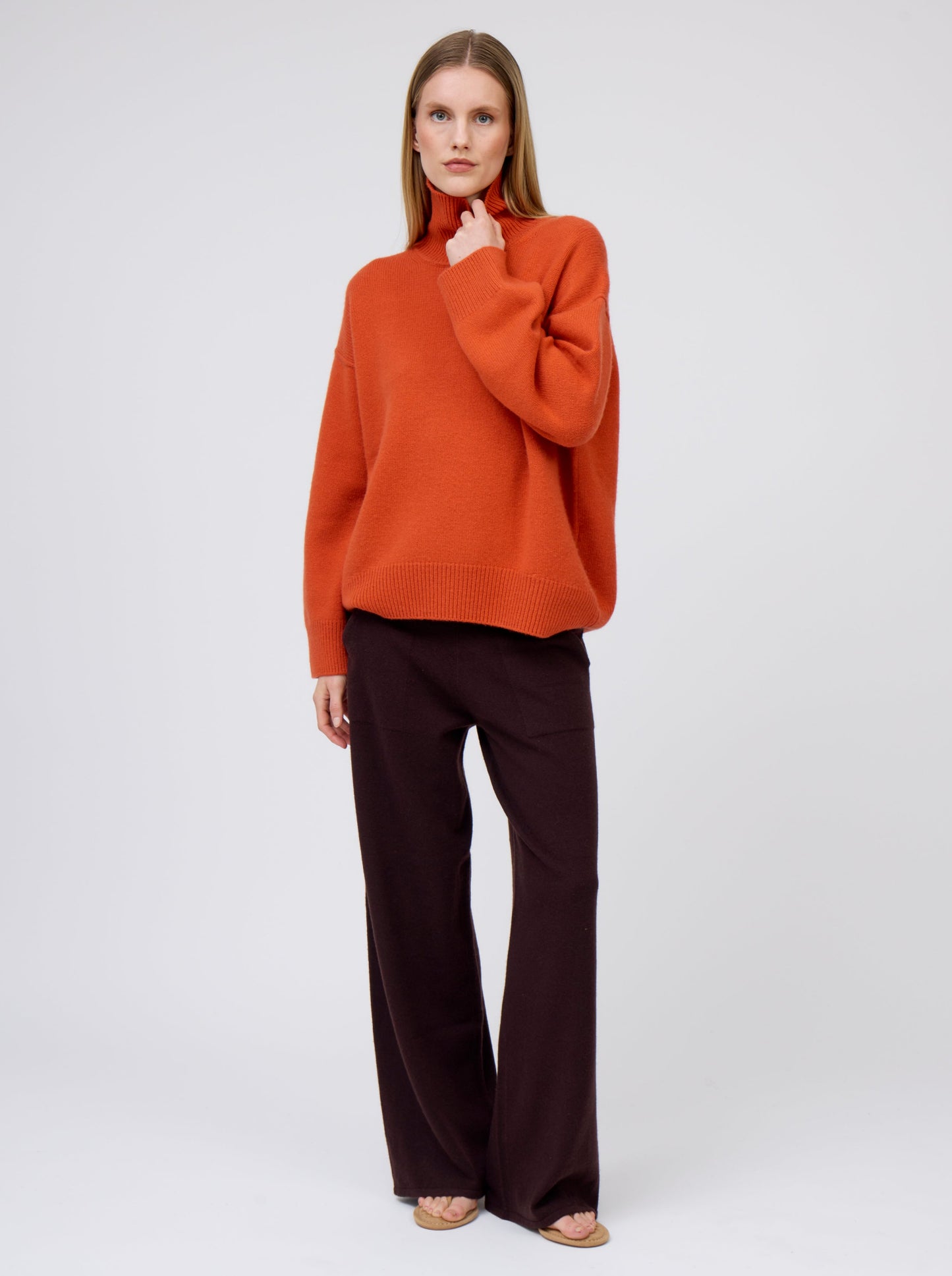 Posed model wears deep orange cashmere high roll neck jumper with wide leg dark brown pants and suede thongs.