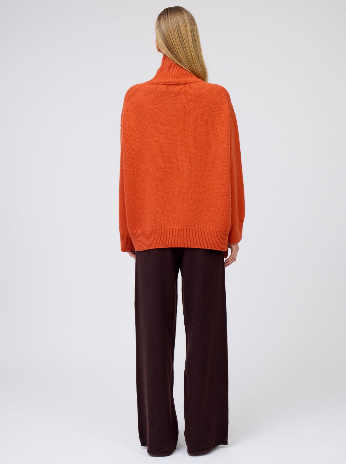 Back view model wears deep orange cashmere roll neck jumper with wide leg dark brown pants.