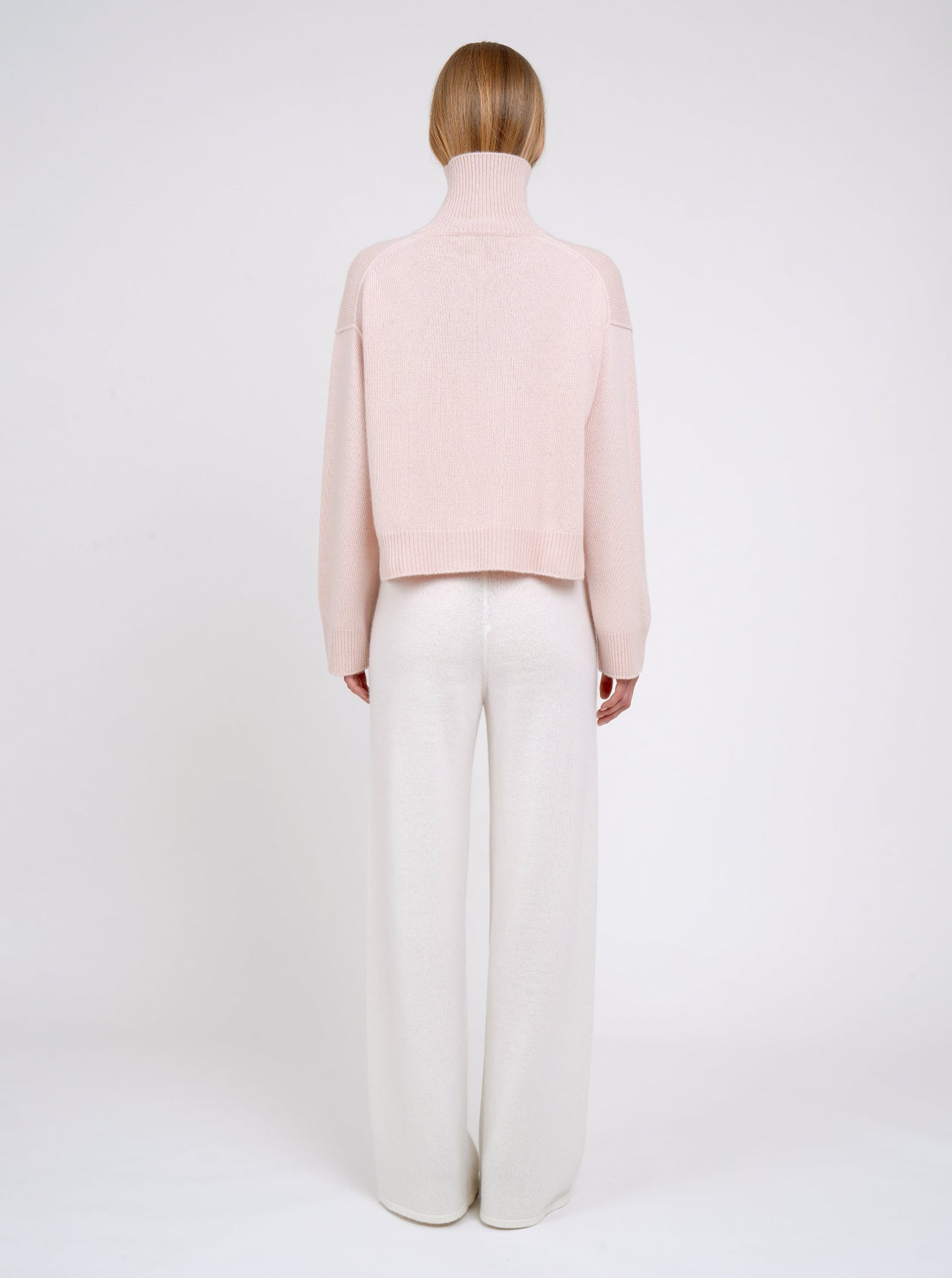 Model wearing a light pink sweater and white pants on a white background