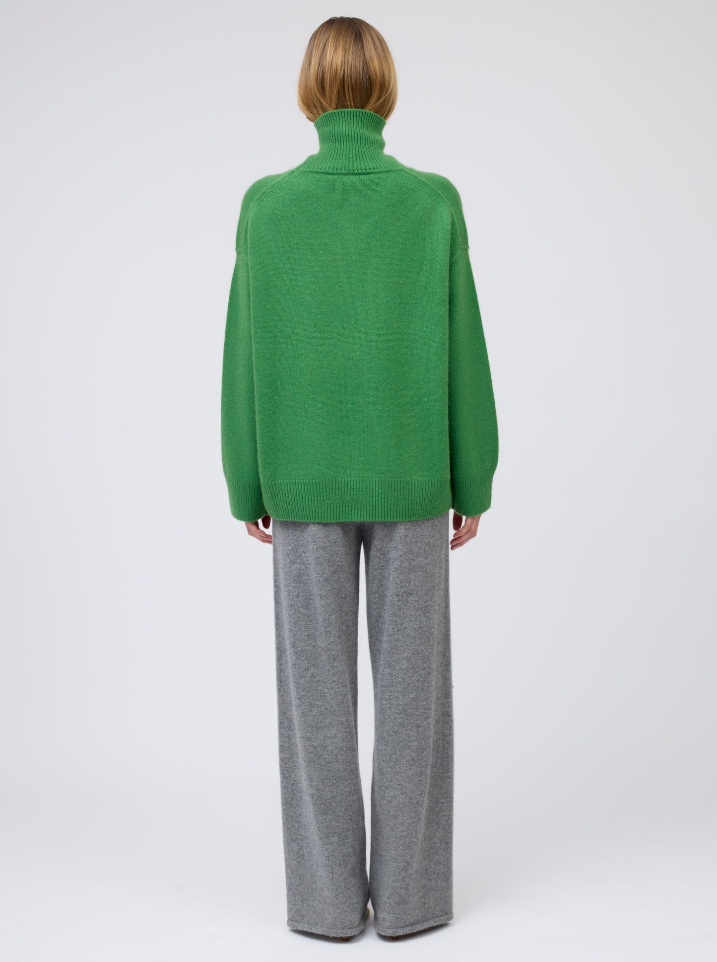 Back view model wears pine green cashmere roll neck oversized jumper with grey cashmere wide leg pants.