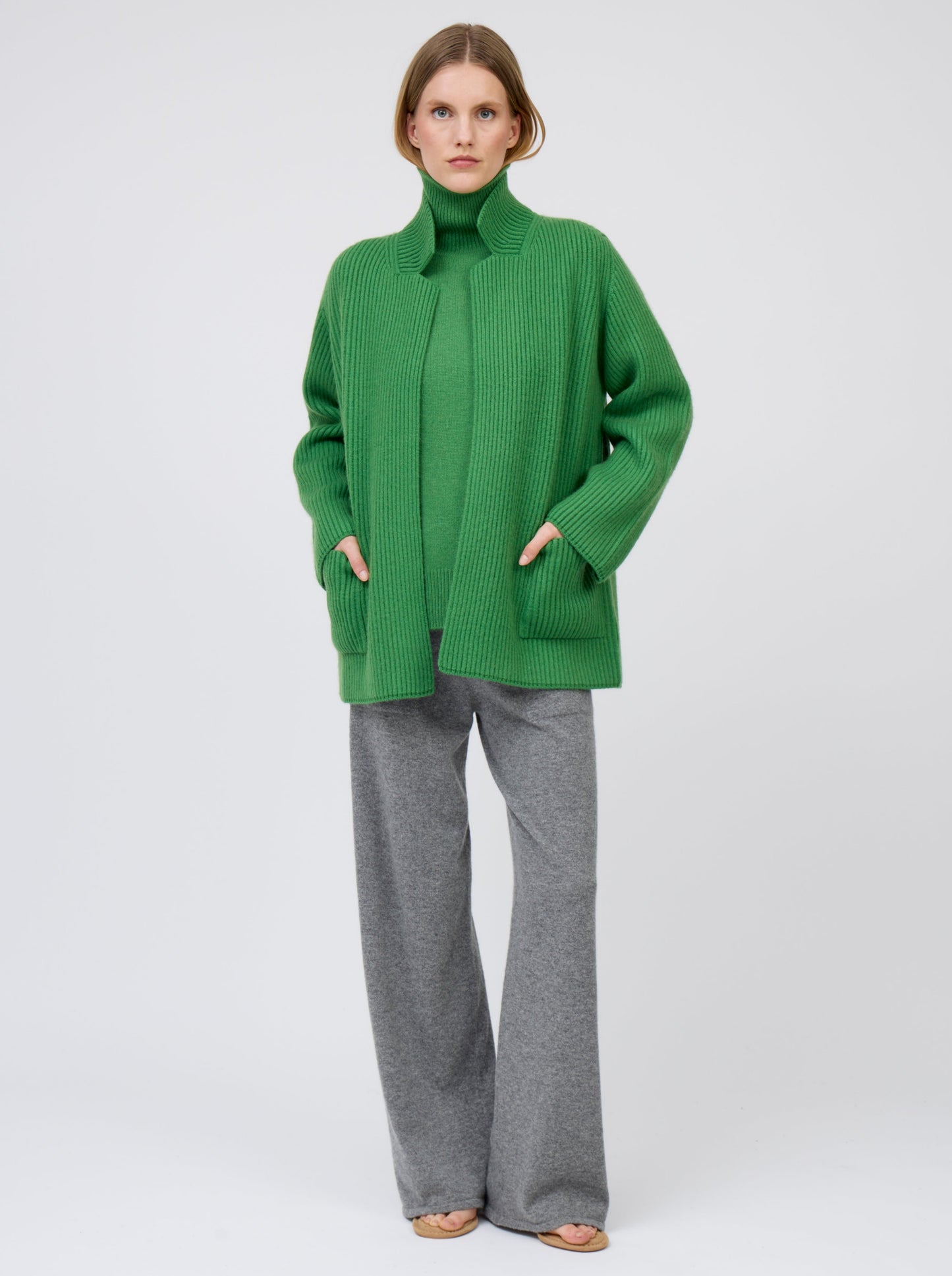 Relaxed, hands in pockets, model wears pine green ribbed knit cashmere hip-length jacket with high collar, wide sleeves and patch pockets; worn with pine green roll neck jumper and grey wide leg pants.