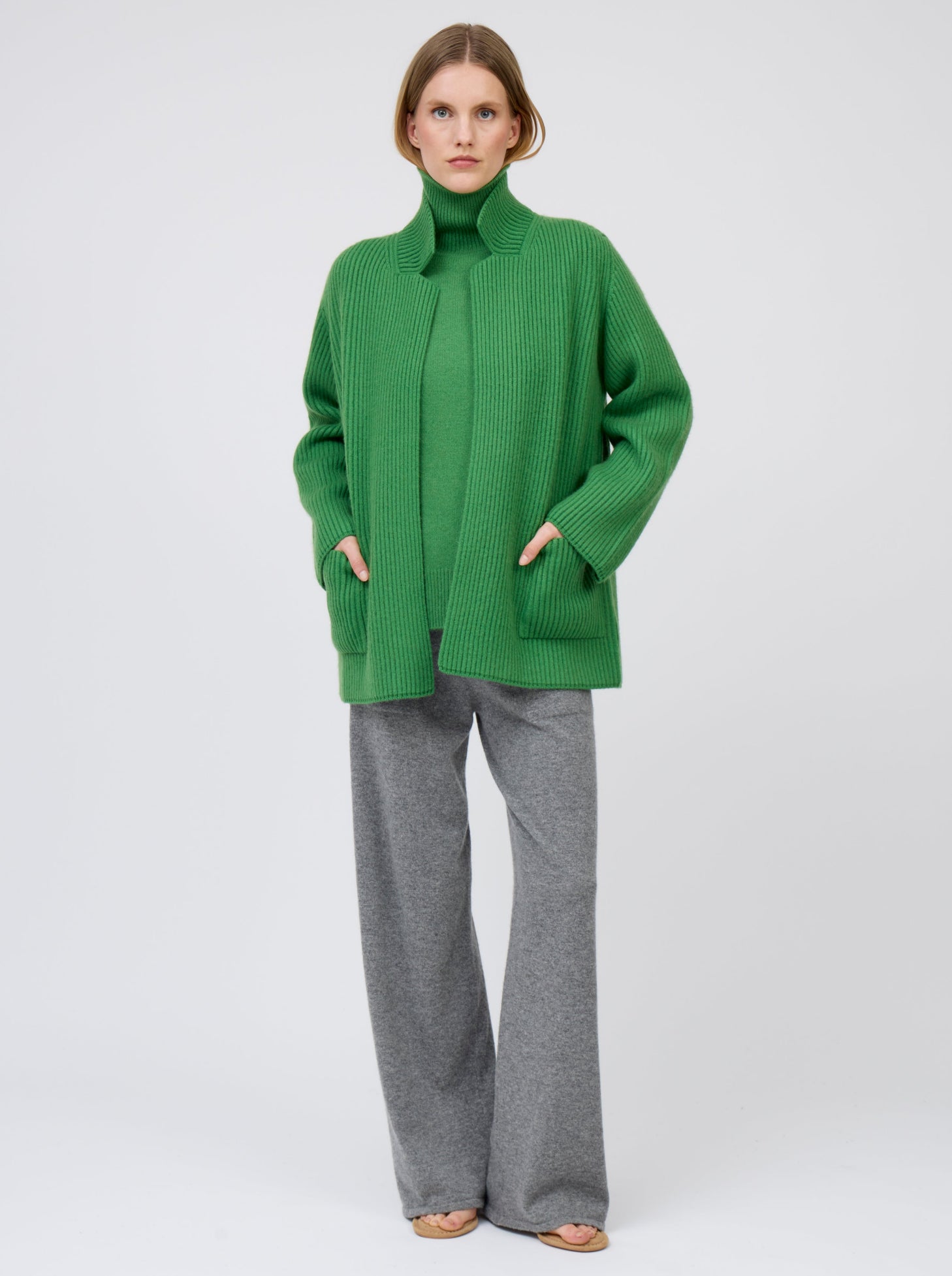 Relaxed, hands in pockets, model wears pine green ribbed knit cashmere hip-length jacket with high collar, wide sleeves and patch pockets; worn with pine green roll neck jumper and grey wide leg pants.