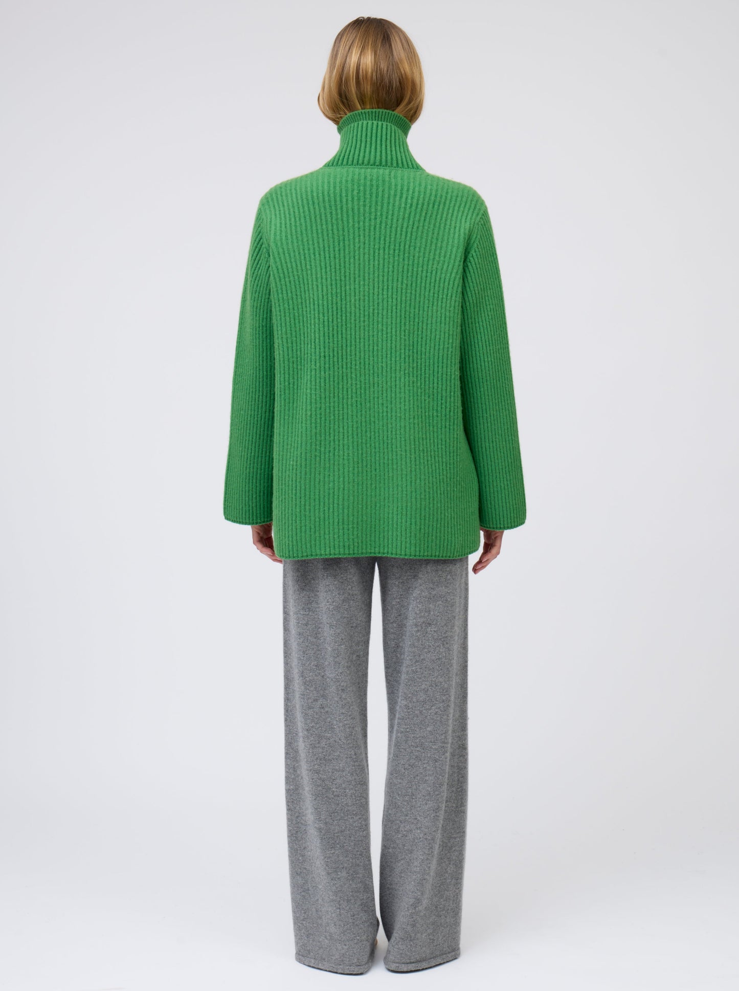 Back view model wears pine green ribbed knit cashmere hip-length jacket with high collar, wide sleeves and patch pockets; worn with grey wide leg pants.