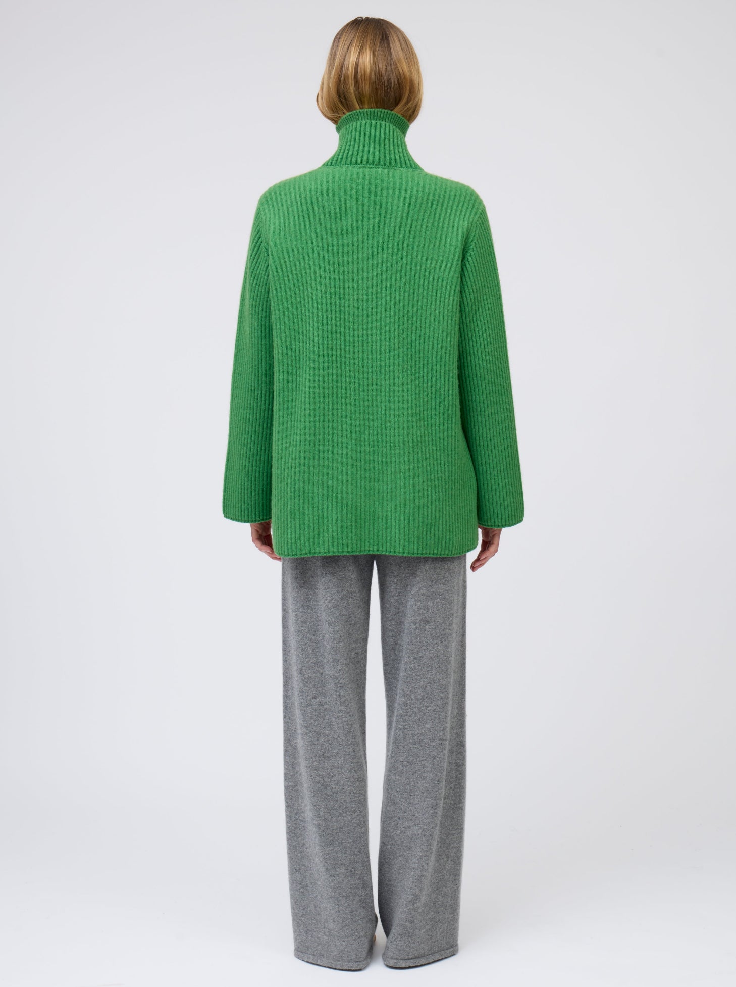 Back view model wears pine green ribbed knit cashmere hip-length jacket with high collar, wide sleeves and patch pockets; worn with grey wide leg pants.