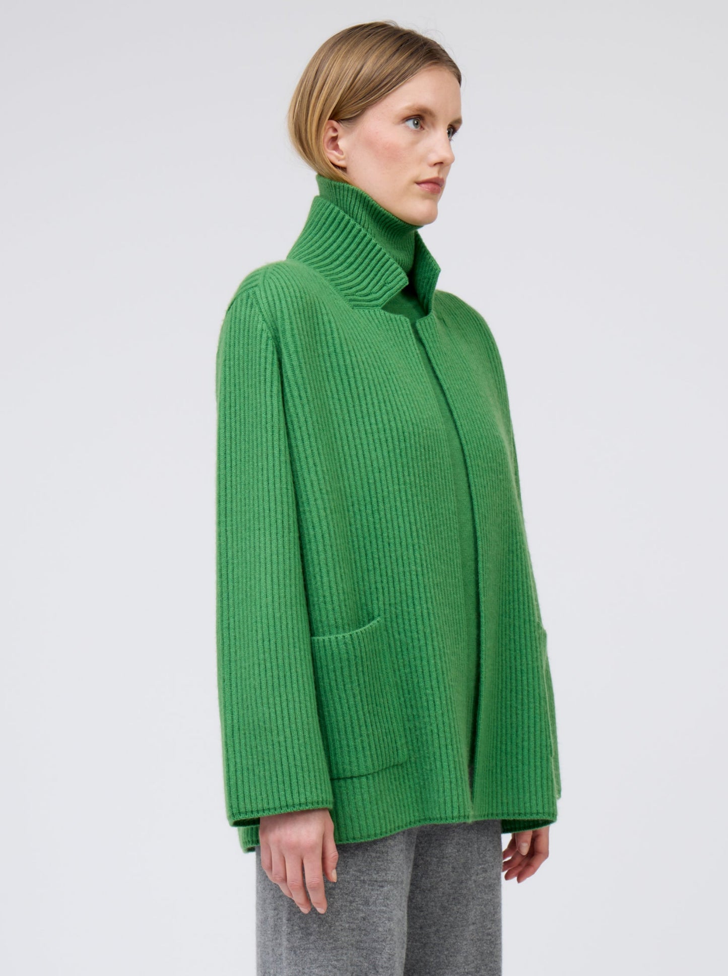 Close-up model wears pine green ribbed knit cashmere hip-length jacket with high collar, wide sleeves and patch pockets; worn with pine green roll neck jumper and grey wide leg pants.