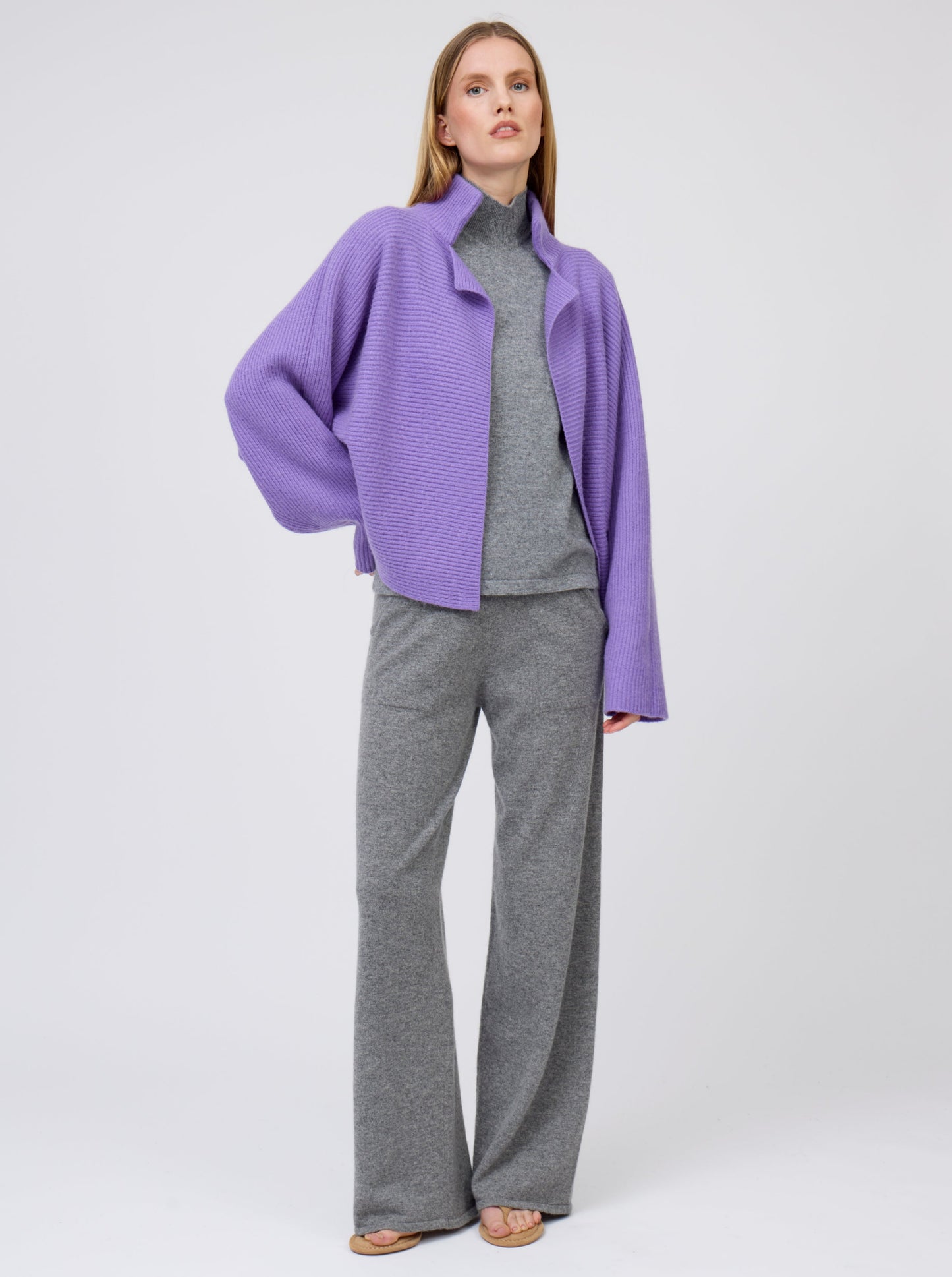 Model, hand on hip, wears purple cashmere ribbed knit jacket with kimono sleeves with grey cashmere turtleneck top and wide leg pants.
