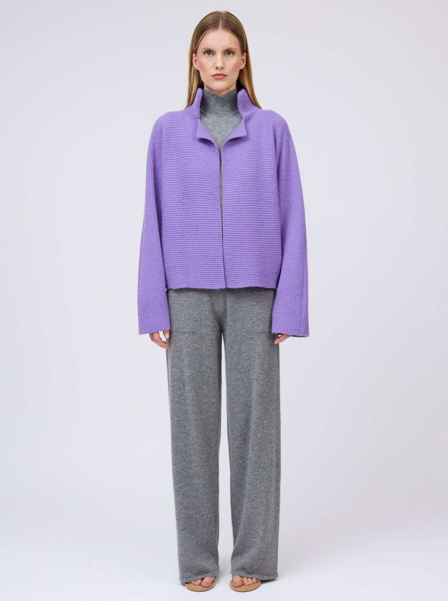 Front view model wears purple cashmere ribbed knit jacket with kimono sleeves and high collar; worn with grey cashmere turtleneck top and wide leg pants.