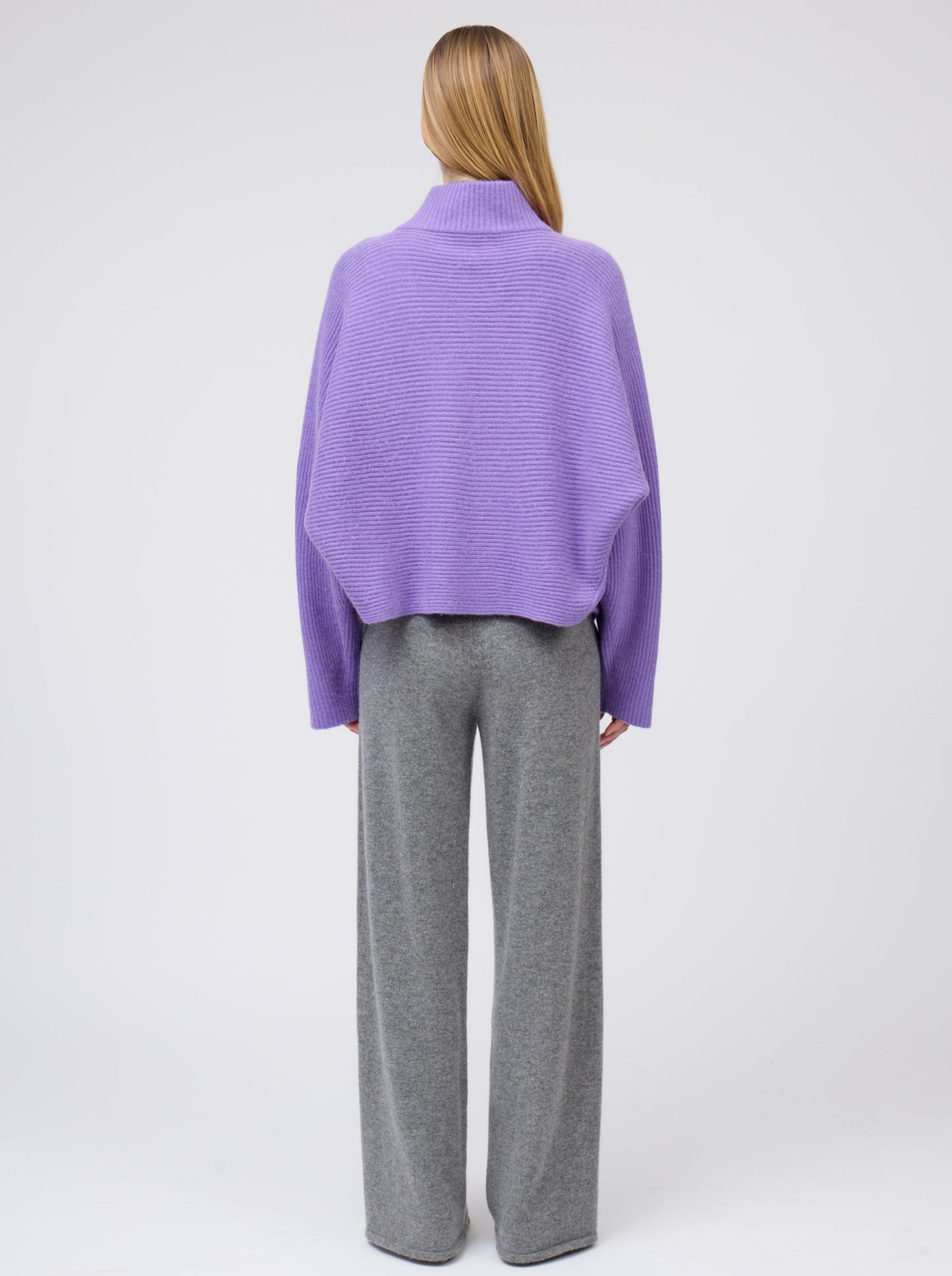 Back view model wears purple cashmere ribbed knit jacket with kimono sleeves and high collar; worn with grey cashmere wide leg pants.