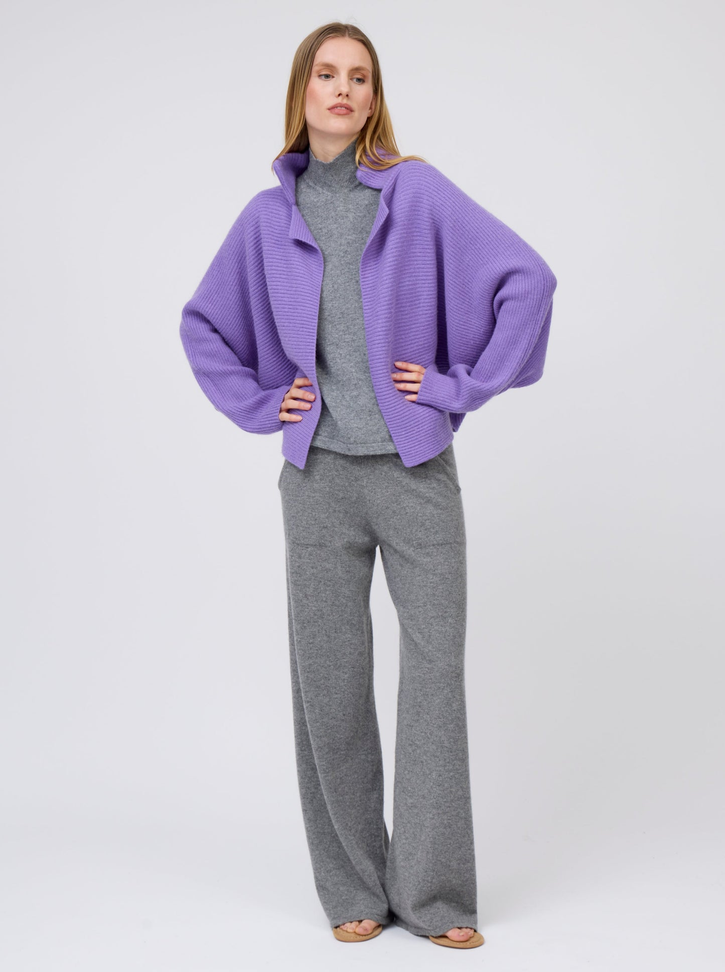 Relaxed pose model wears purple cashmere ribbed knit jacket with kimono sleeves and high collar; worn with grey cashmere turtleneck top and wide leg pants.