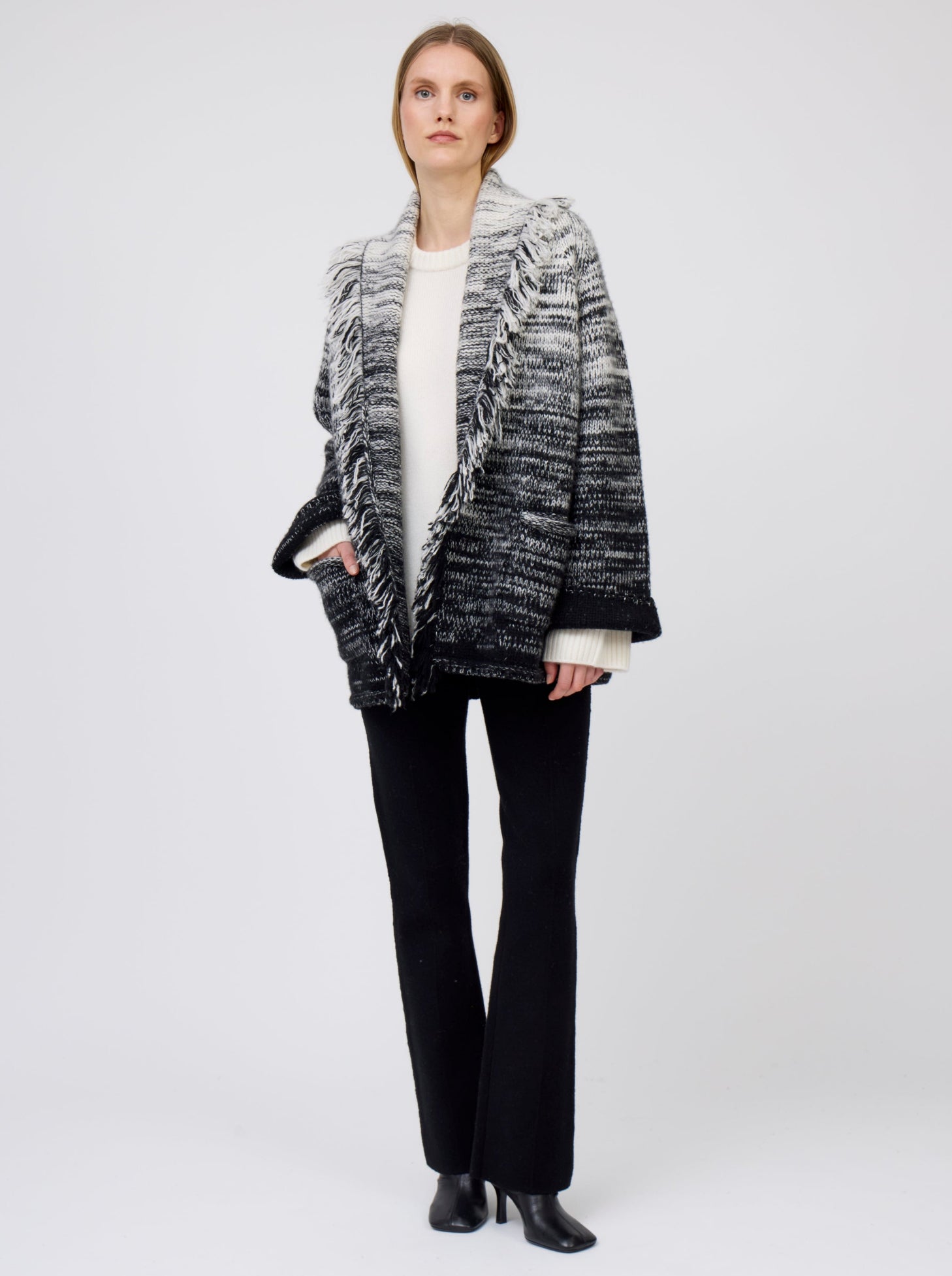 Model wears  gradient colour mix knitted cashmere jacket with shawl collar and pockets, in white and black over white cashmere sweater and  black suit pants.