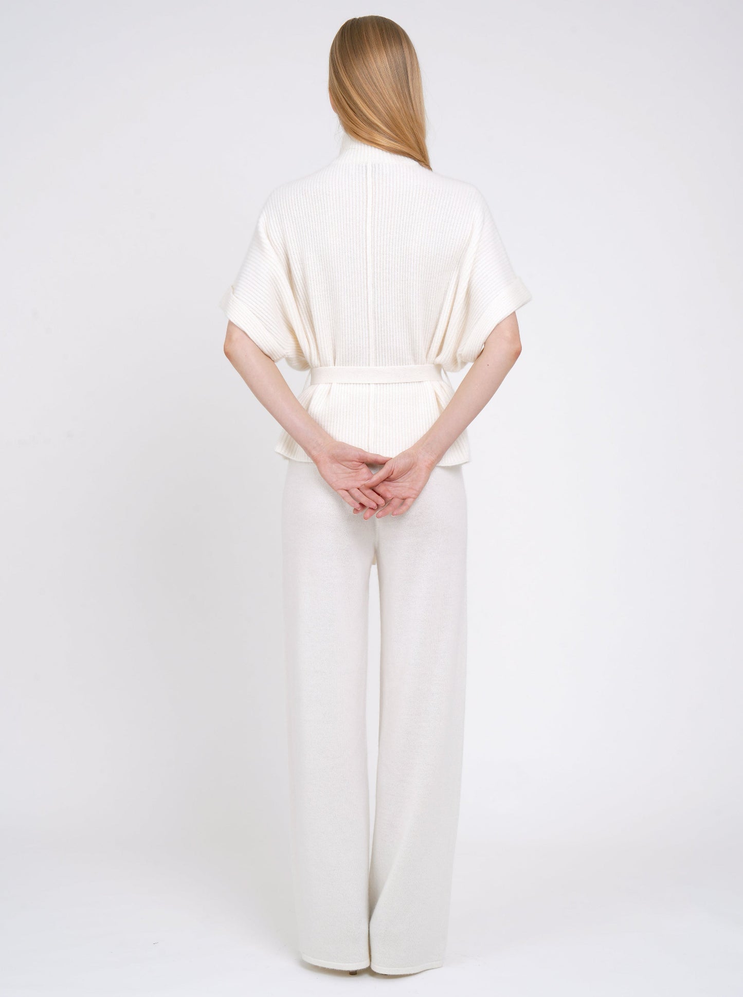 Back view model in white ribbed knit cashmere belted jacket with wide short sleeves and white wide leg knit pants.